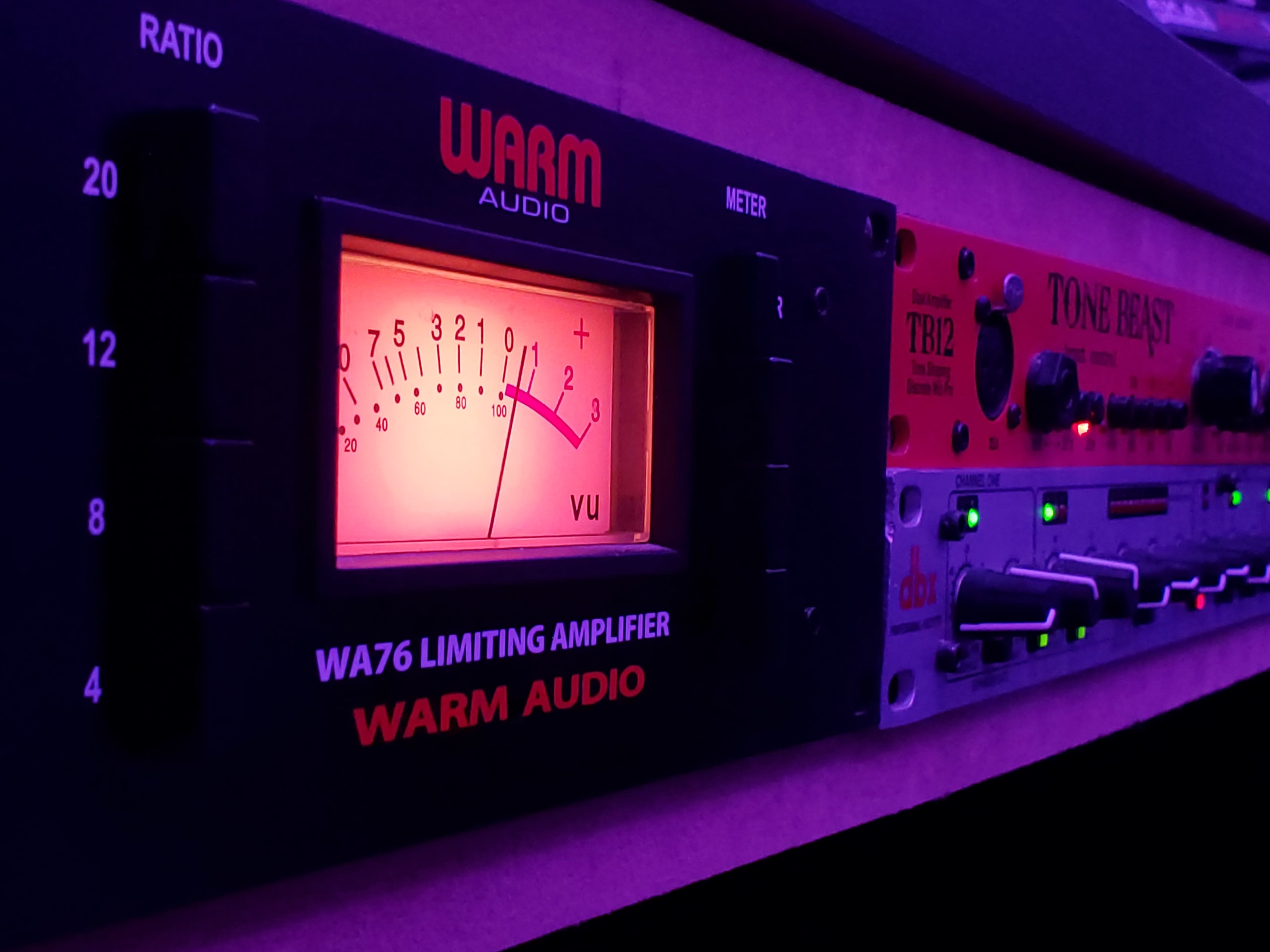 The image shows a Warm Audio WA76 limiting amplifier with a glowing VU meter positioned next to a Tone Beast TB12 audio processor.