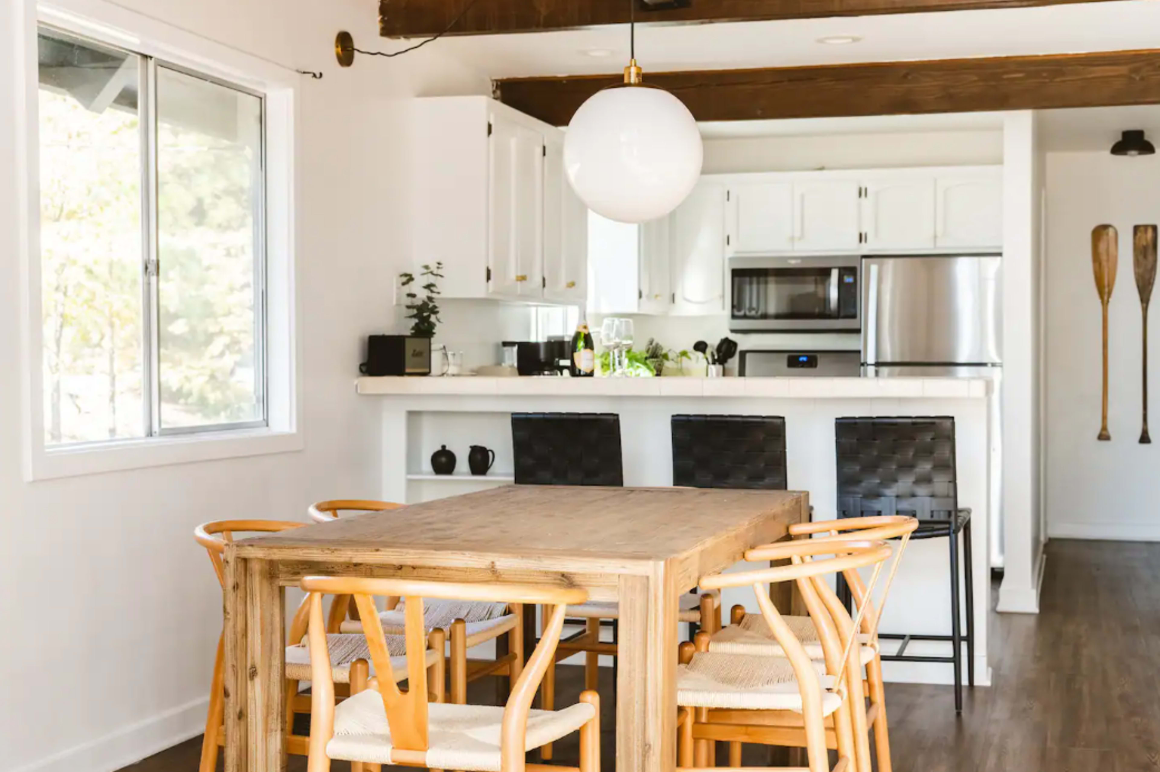 A wooden dining table with chairs is positioned in a bright kitchen area featuring white cabinets and stainless steel appliances.