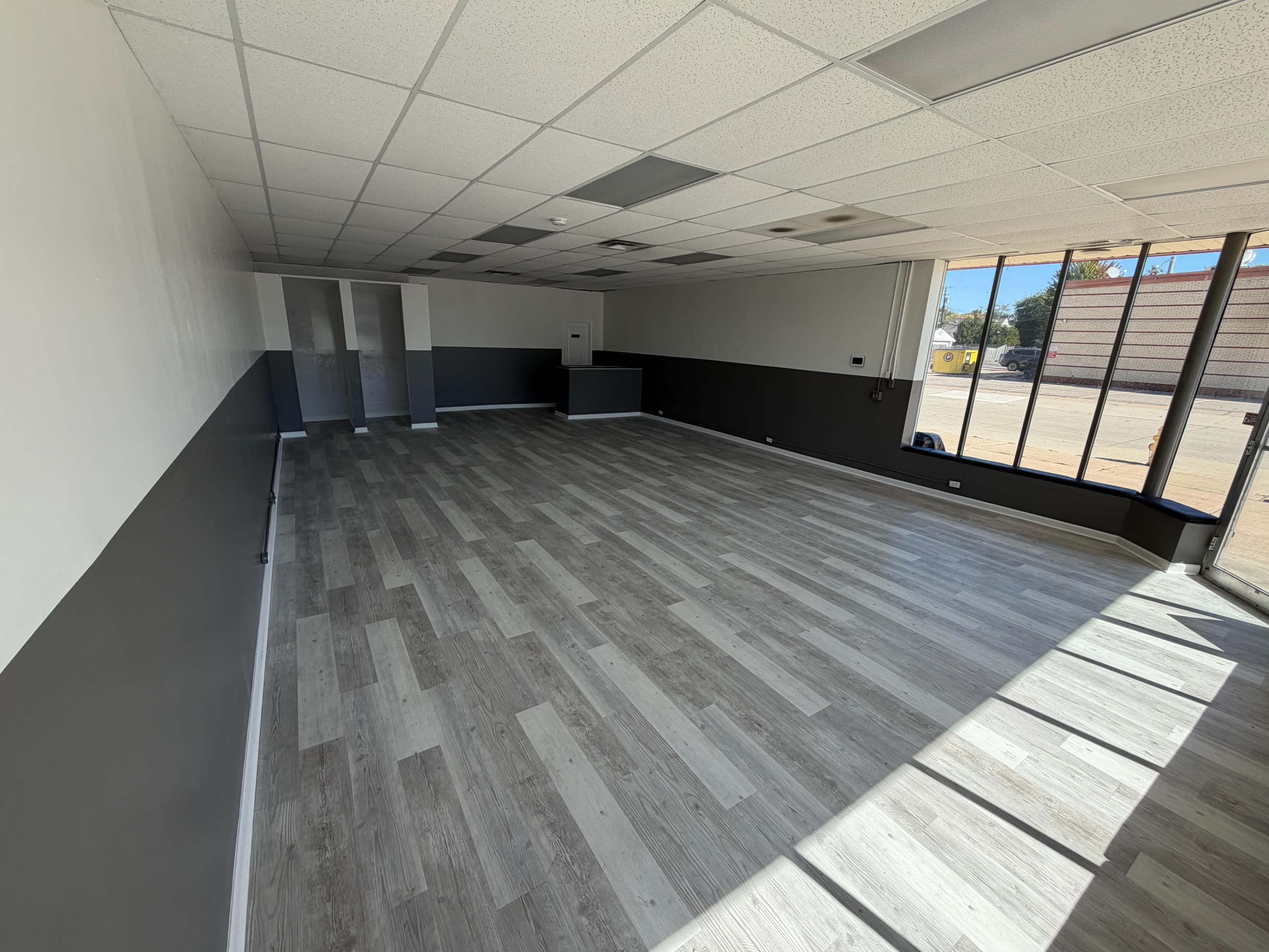 The image shows a spacious, empty room with new gray and beige flooring, white walls, and large windows letting in natural light.