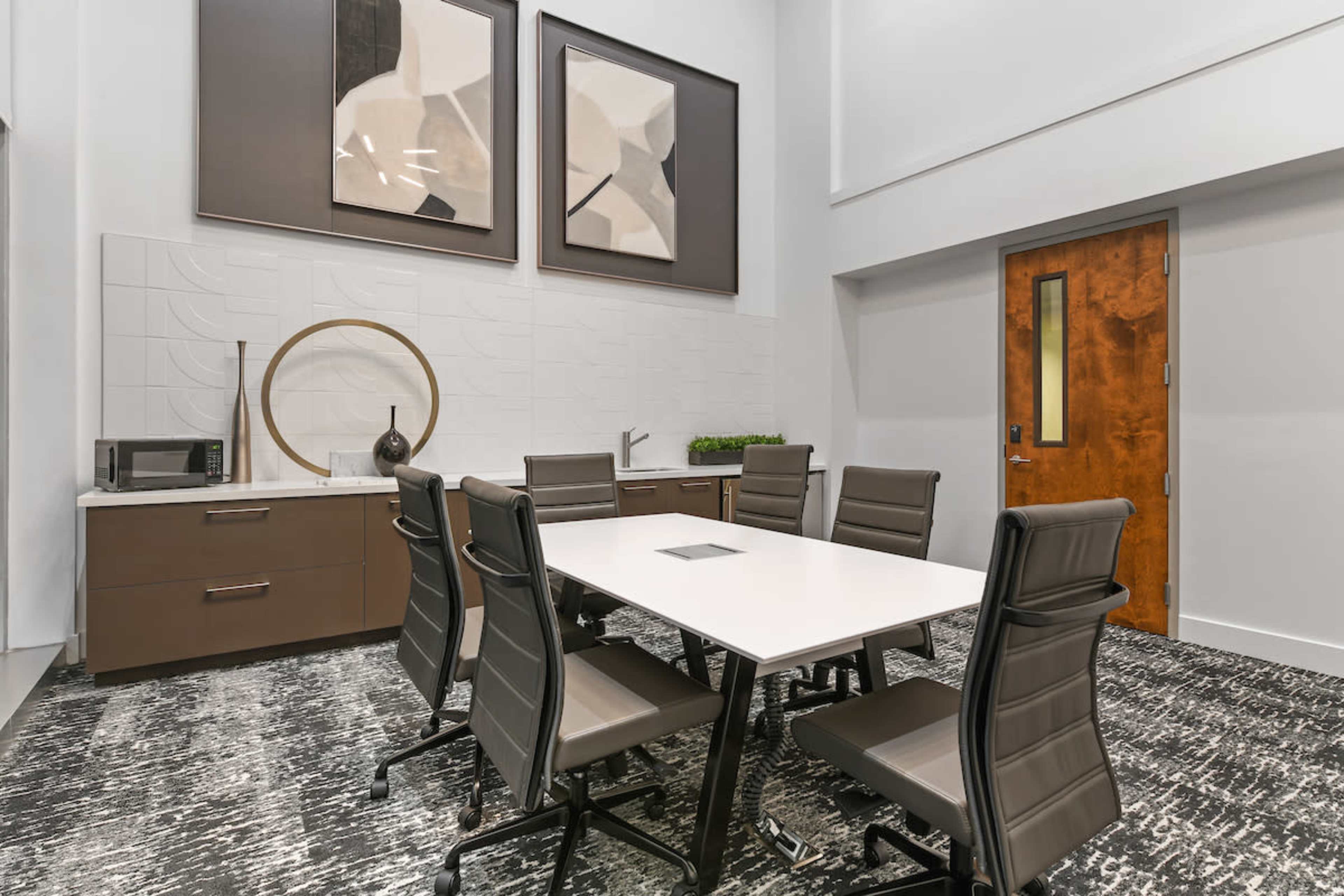A modern conference room features a white table surrounded by six ergonomic chairs, complemented by kitchen amenities and contemporary artwork on the walls.