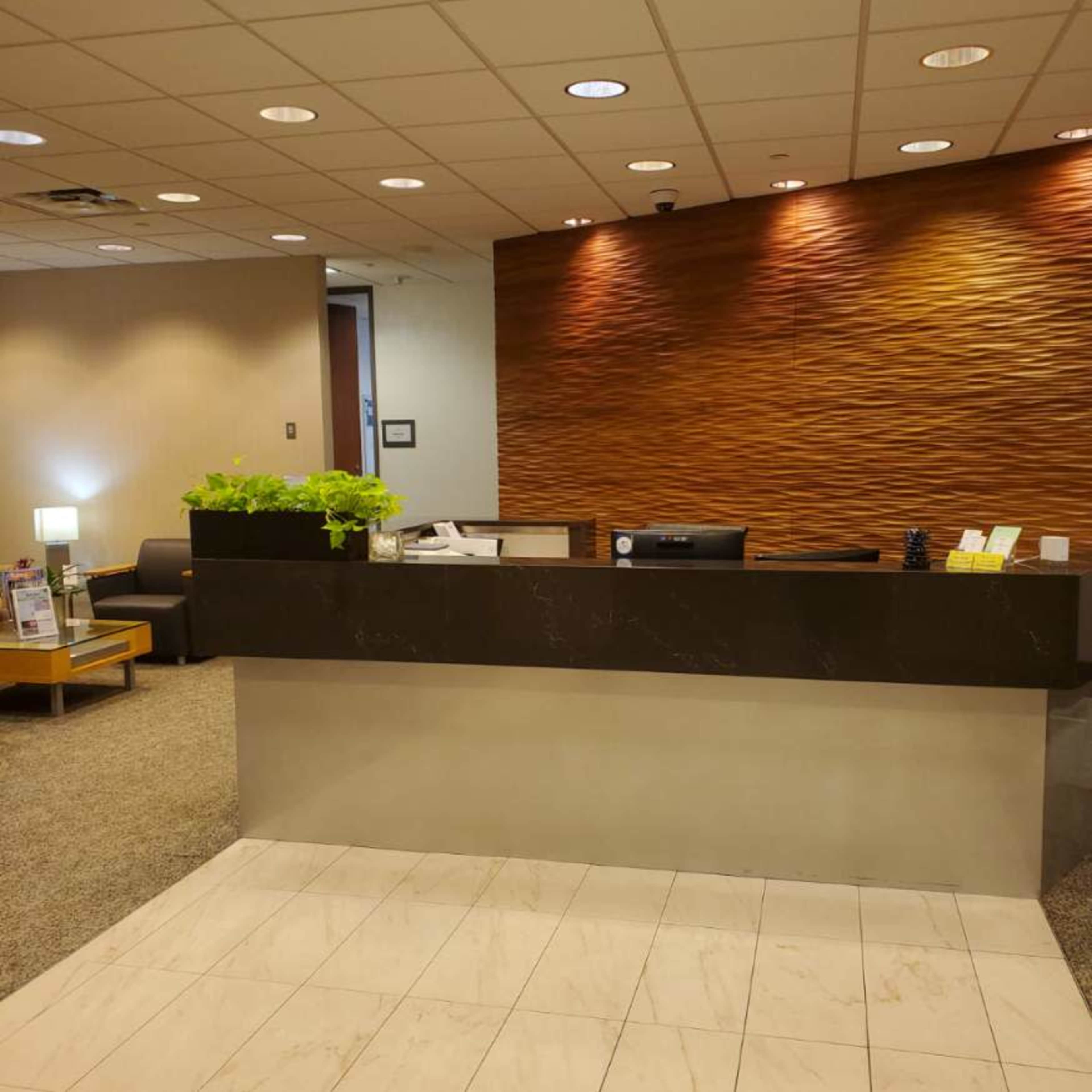A modern reception desk with a dark surface and a green plant atop, set against a textured, warm-toned wall in a well-lit lobby area.