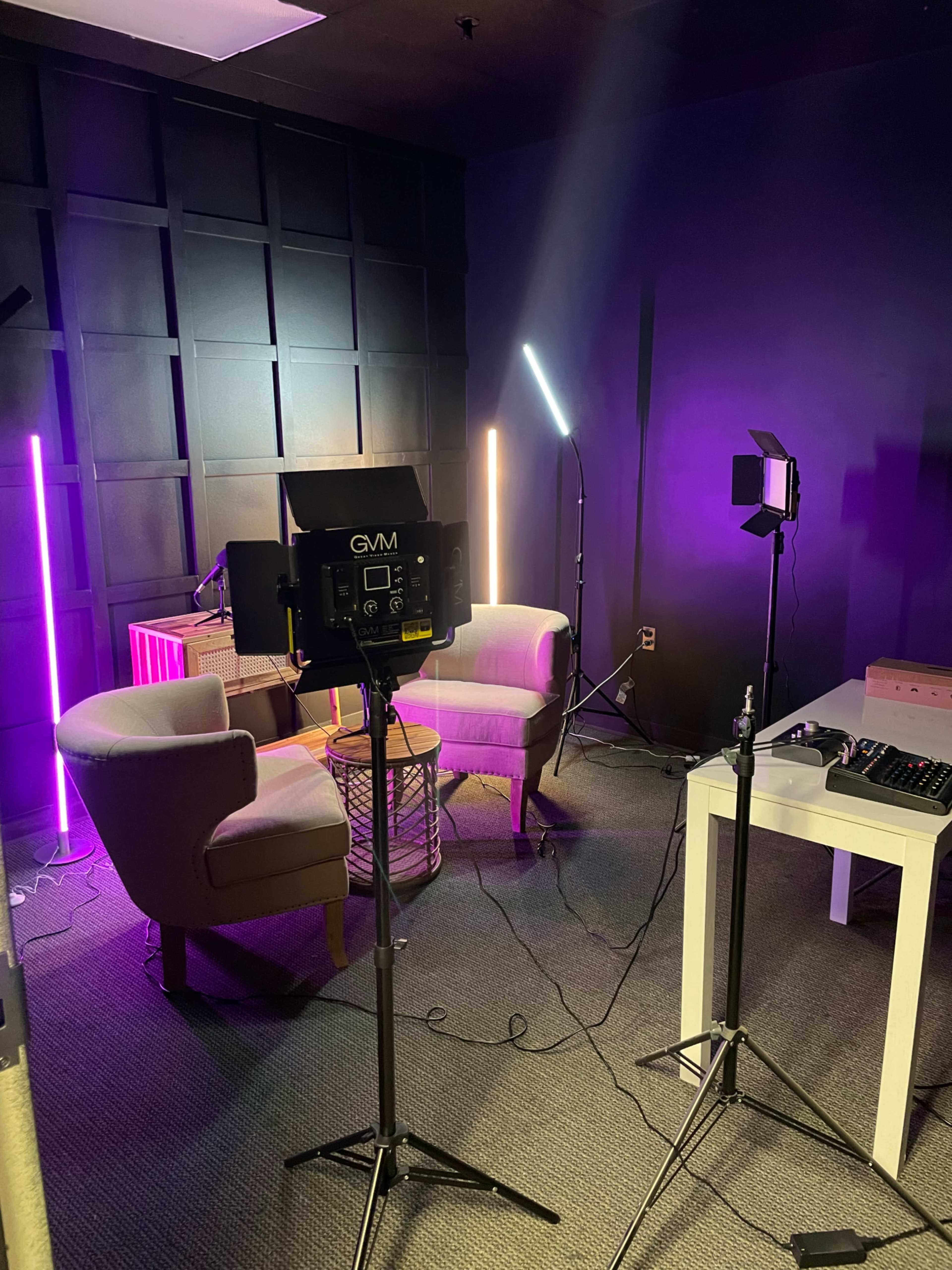 A small studio setup with two chairs, several lights positioned on stands, and a table with equipment in a room with dark walls.