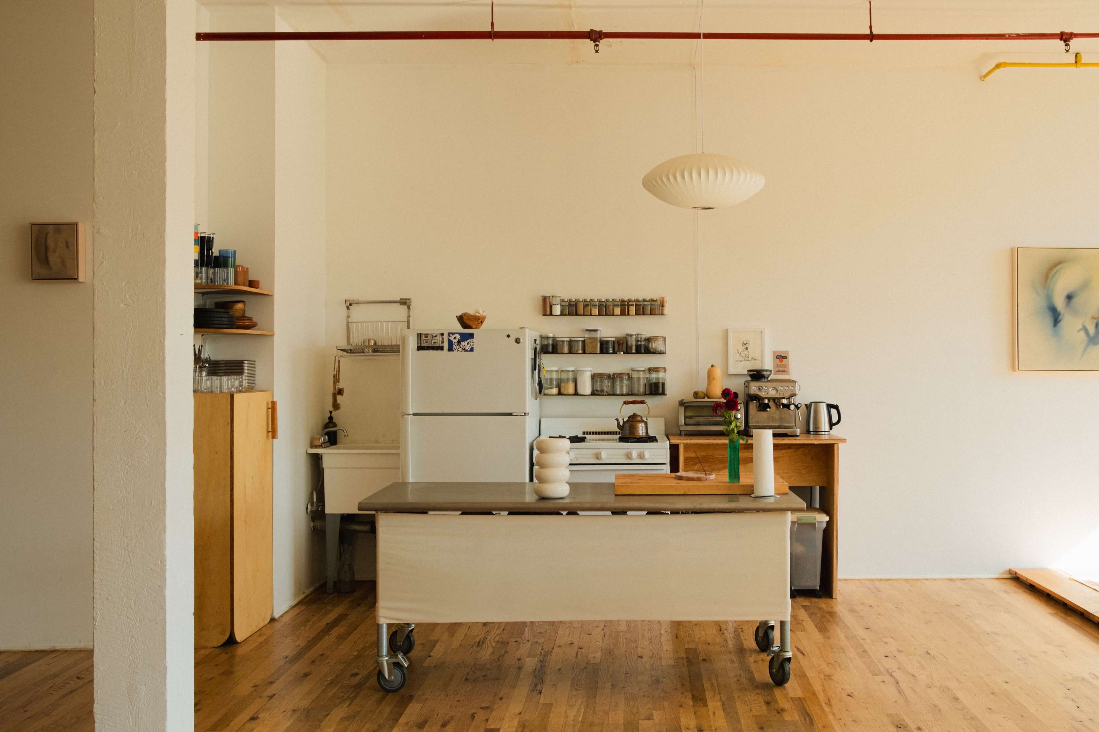 The image shows a spacious, modern kitchen with a central island on wheels, white appliances, shelves filled with jars, and a minimalistic decor.