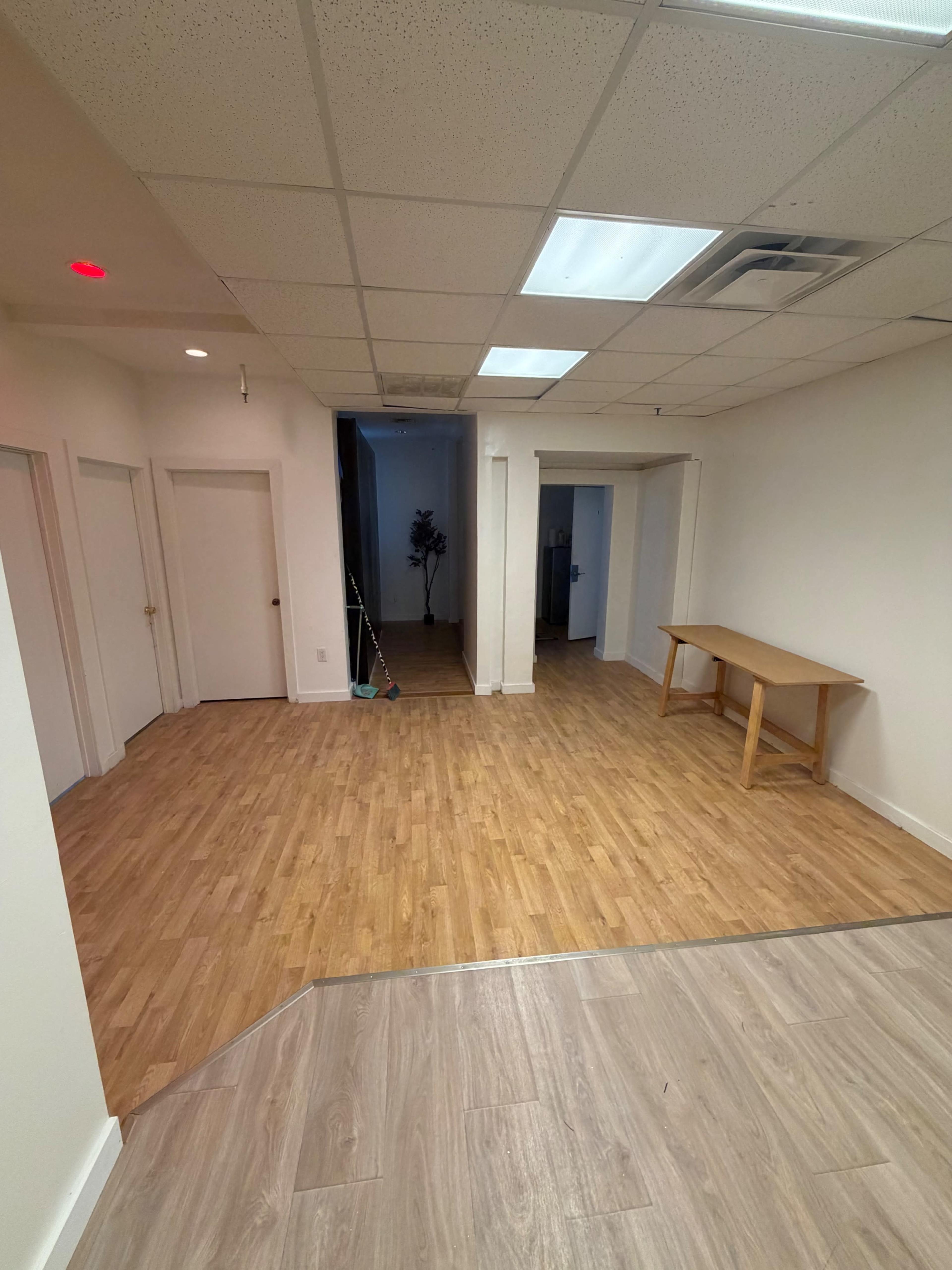 The image shows a spacious, empty room with wooden flooring, featuring doorways on the left and a table against the wall on the right.