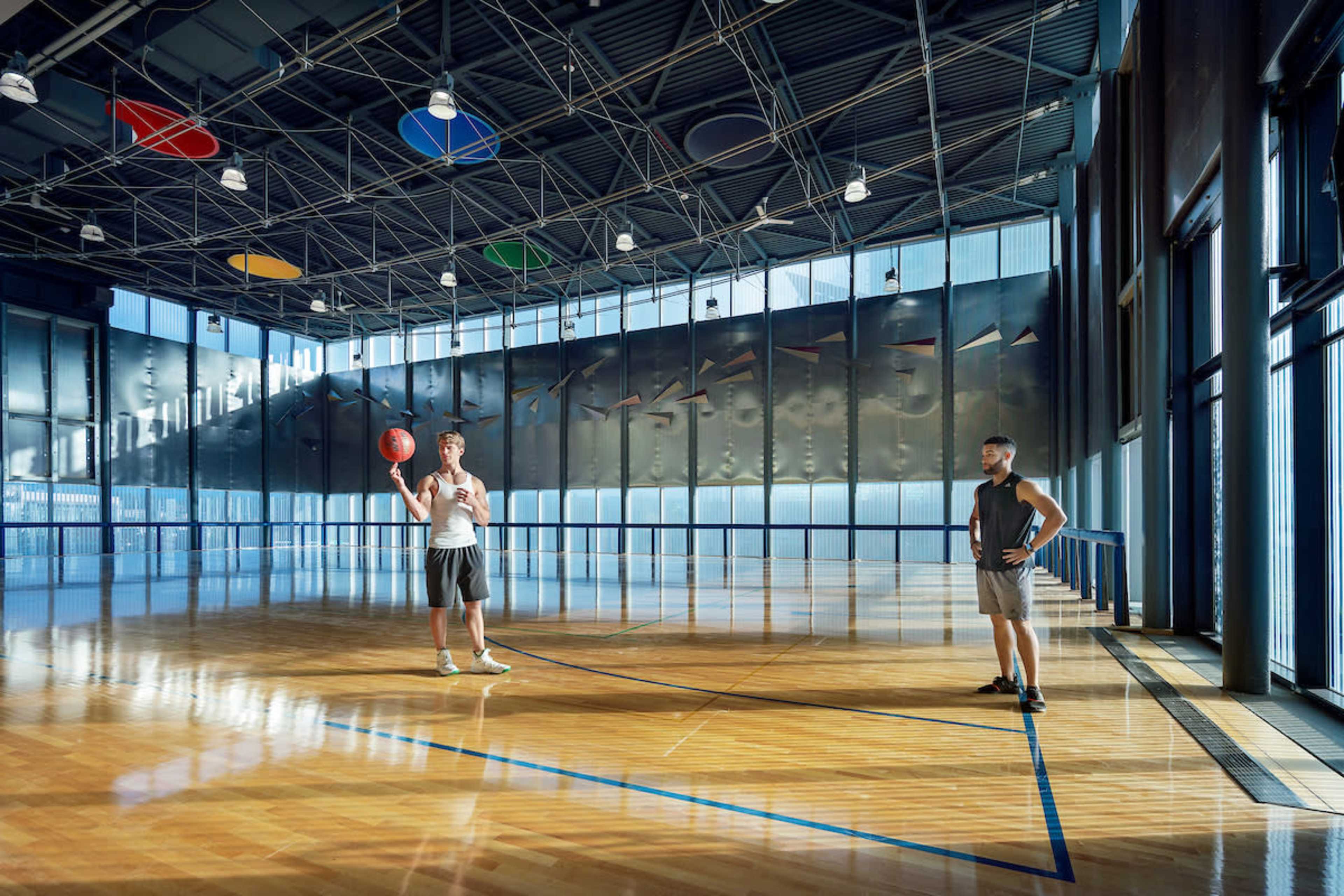 Two men stand on a polished wooden basketball court, one holding a basketball, in a brightly lit indoor sports facility.