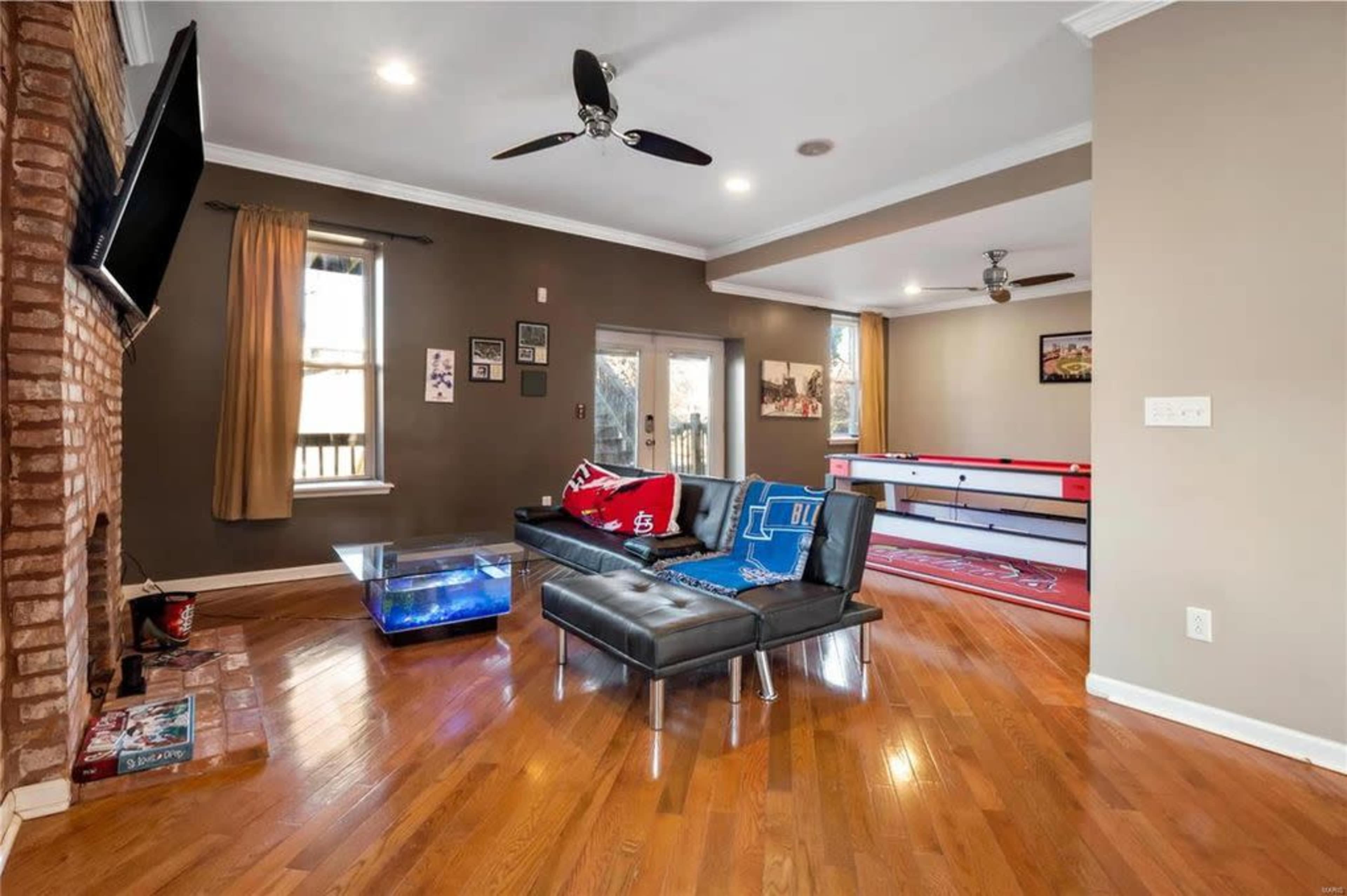 A living room featuring a black leather sectional sofa, a mounted flat-screen TV, and a wooden floor with a coffee table and a gaming area in the background.
