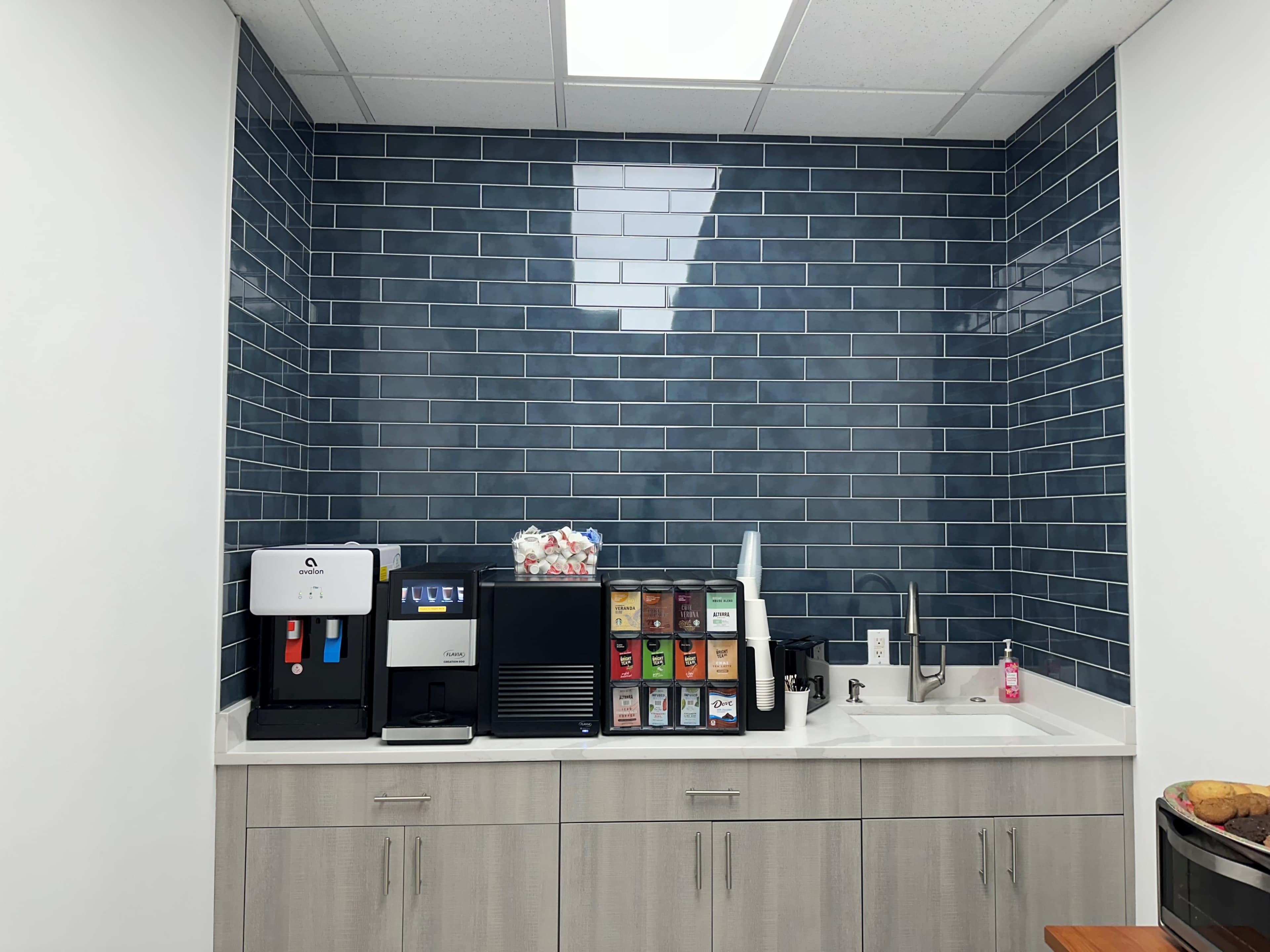 The image shows a kitchenette area featuring a countertop with a coffee machine, a beverage dispenser, and a sink against a blue tiled wall.