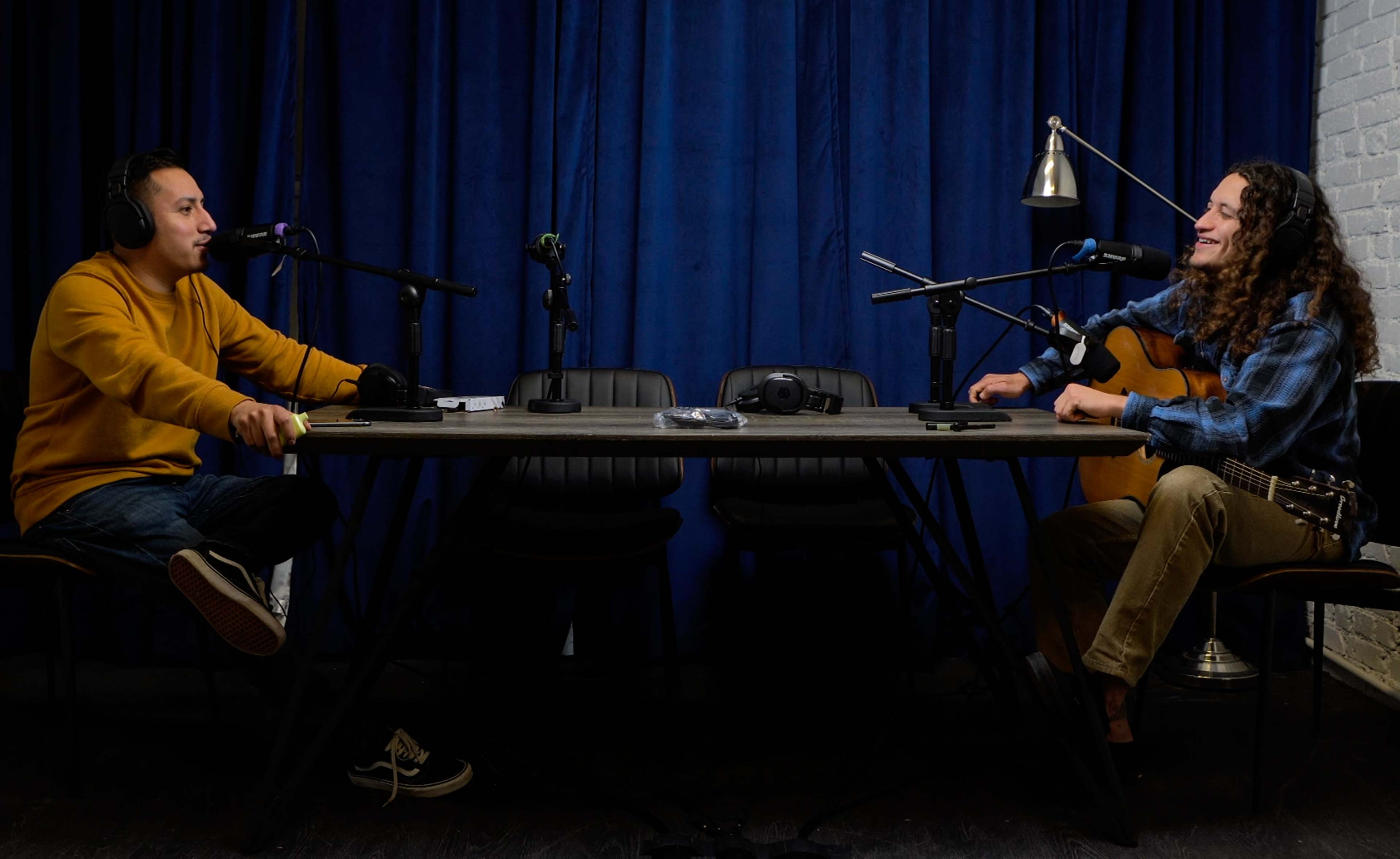 Two individuals sit at a table in a sound studio, equipped with microphones and instruments, engaged in a conversation.