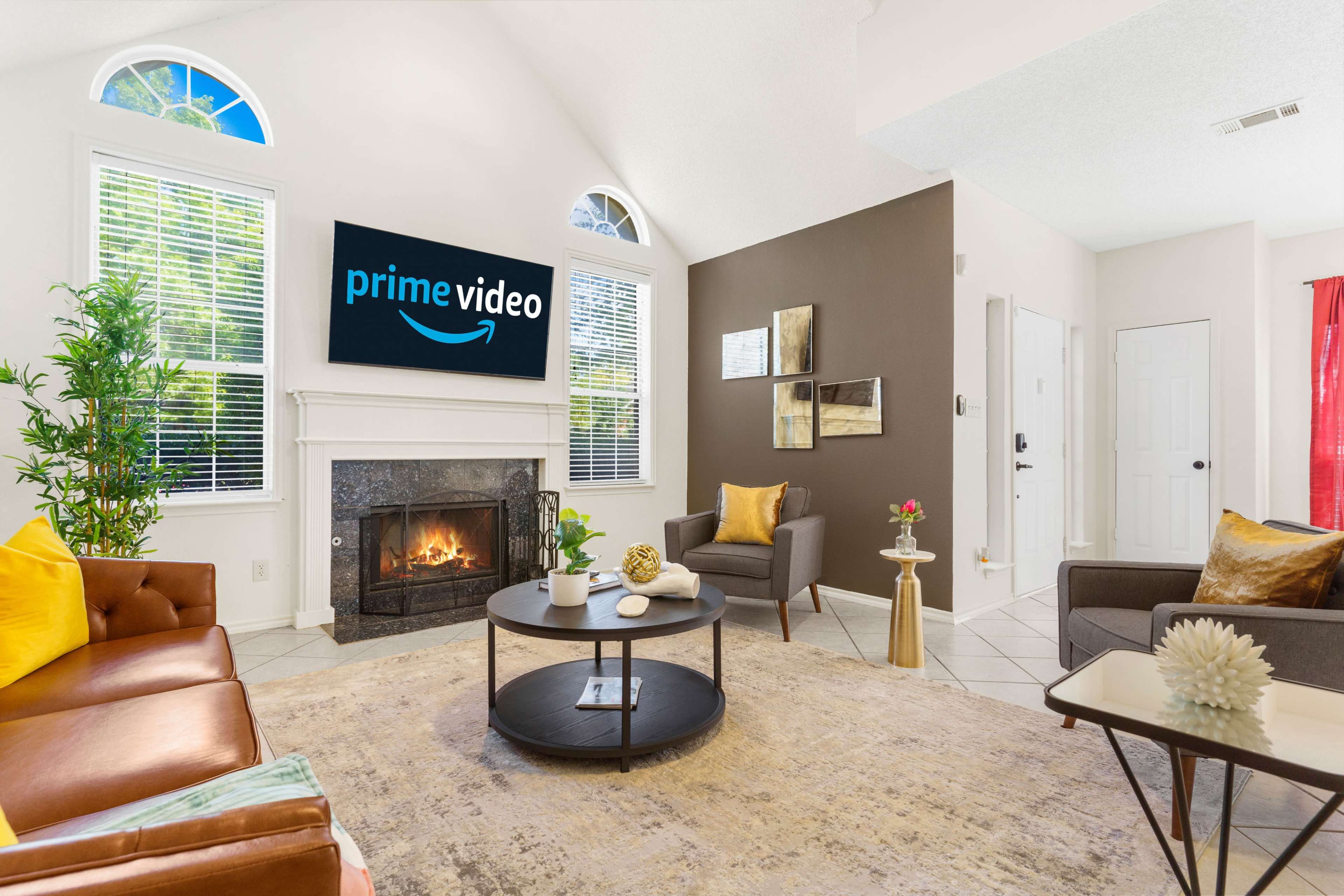 A cozy living room features a leather sofa and armchairs surrounding a coffee table, with a fireplace and a wall-mounted television displaying the Prime Video logo.