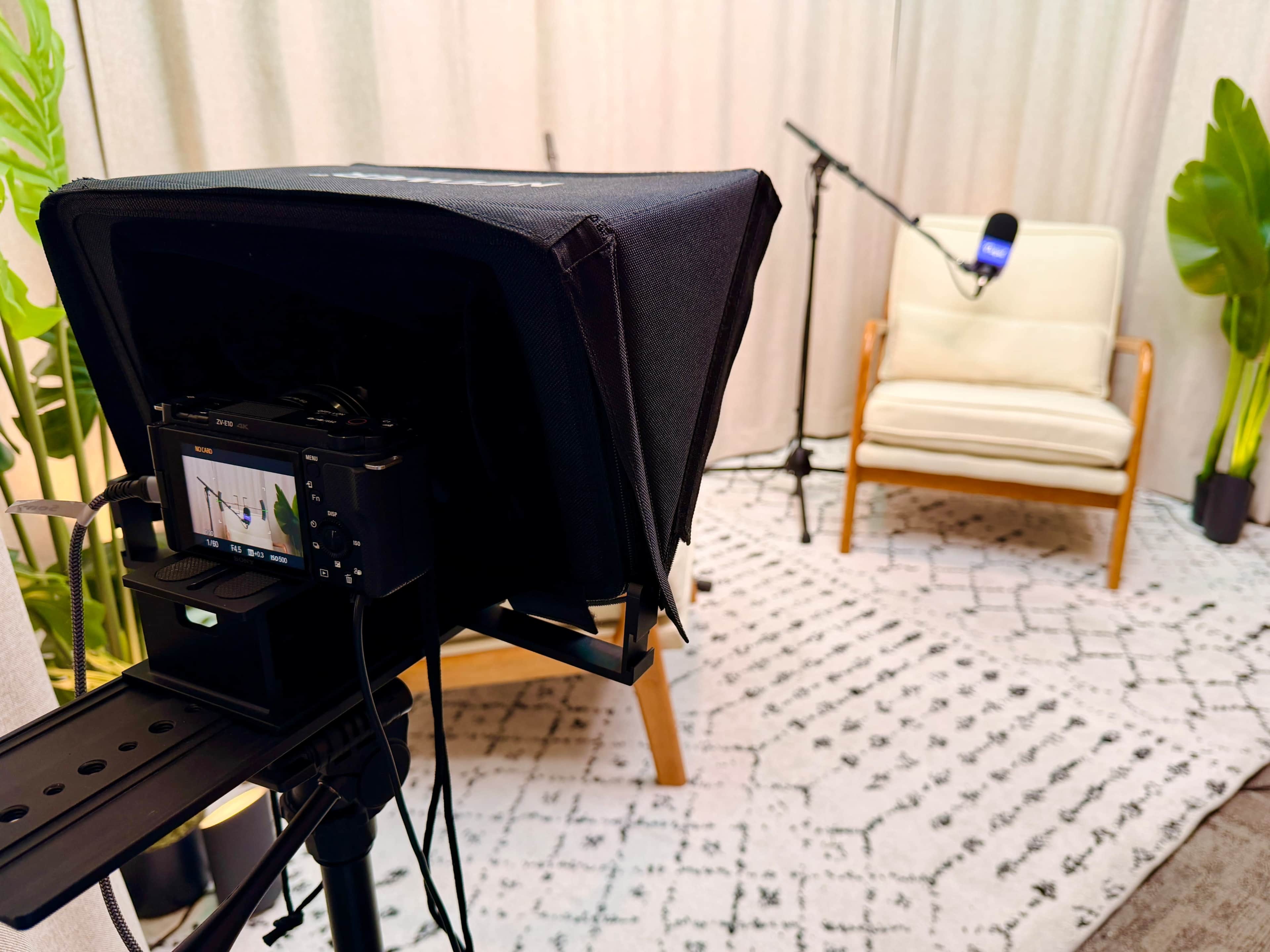The image shows a video recording setup featuring a camera with a teleprompter facing a vacant chair in a well-lit room decorated with plants and a microphone on a stand.