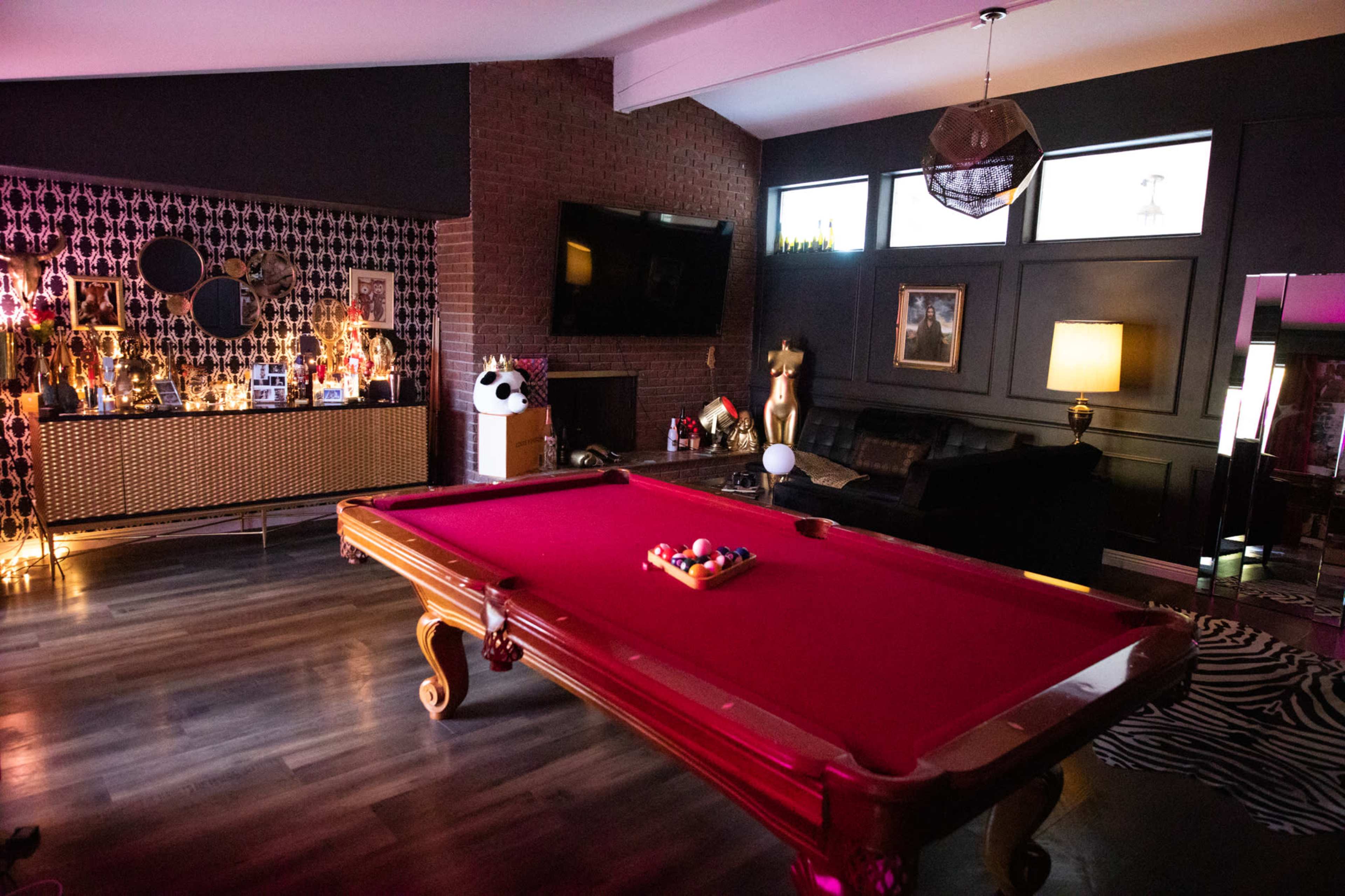 A billiard table with a red felt surface is centered in a stylish game room that features decorative elements like a panda figure and a gold statue.