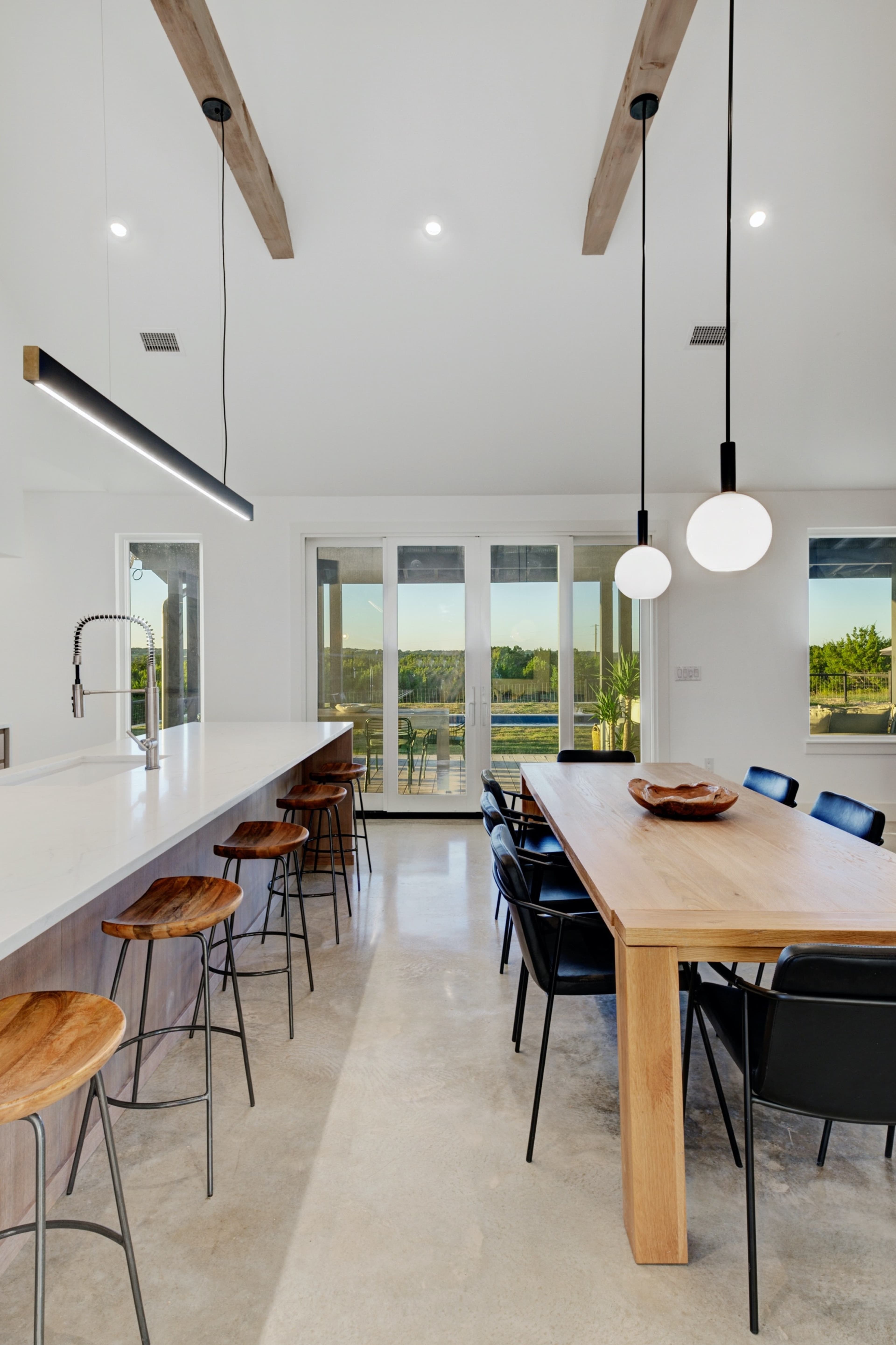 A modern kitchen and dining area with a long wooden table, black chairs, a white countertop, and large windows overlooking a view.