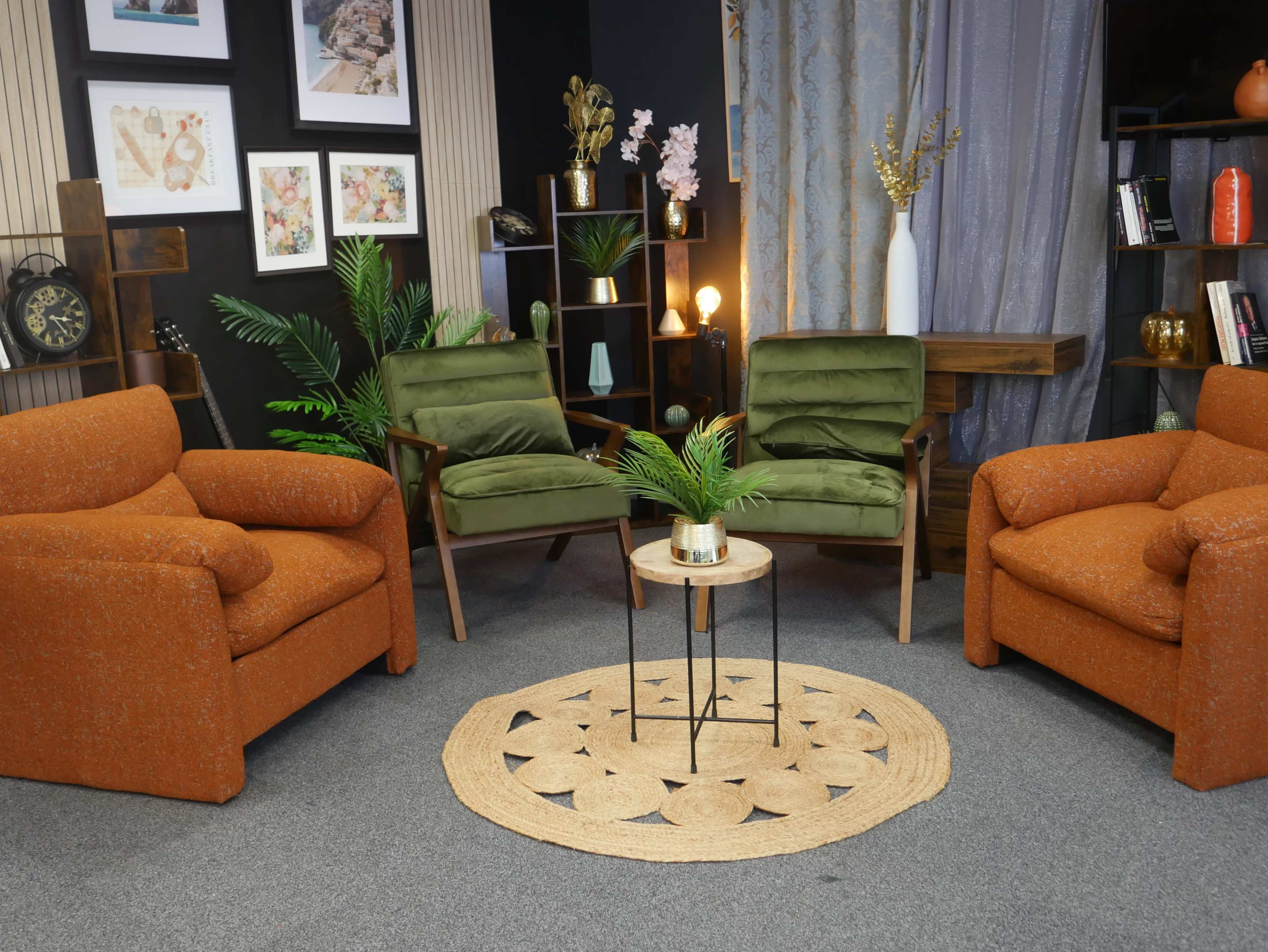 A cozy interior setup features three armchairs—two orange and one green—arranged around a small circular table on a patterned rug, with decorative plants and framed art on the walls.