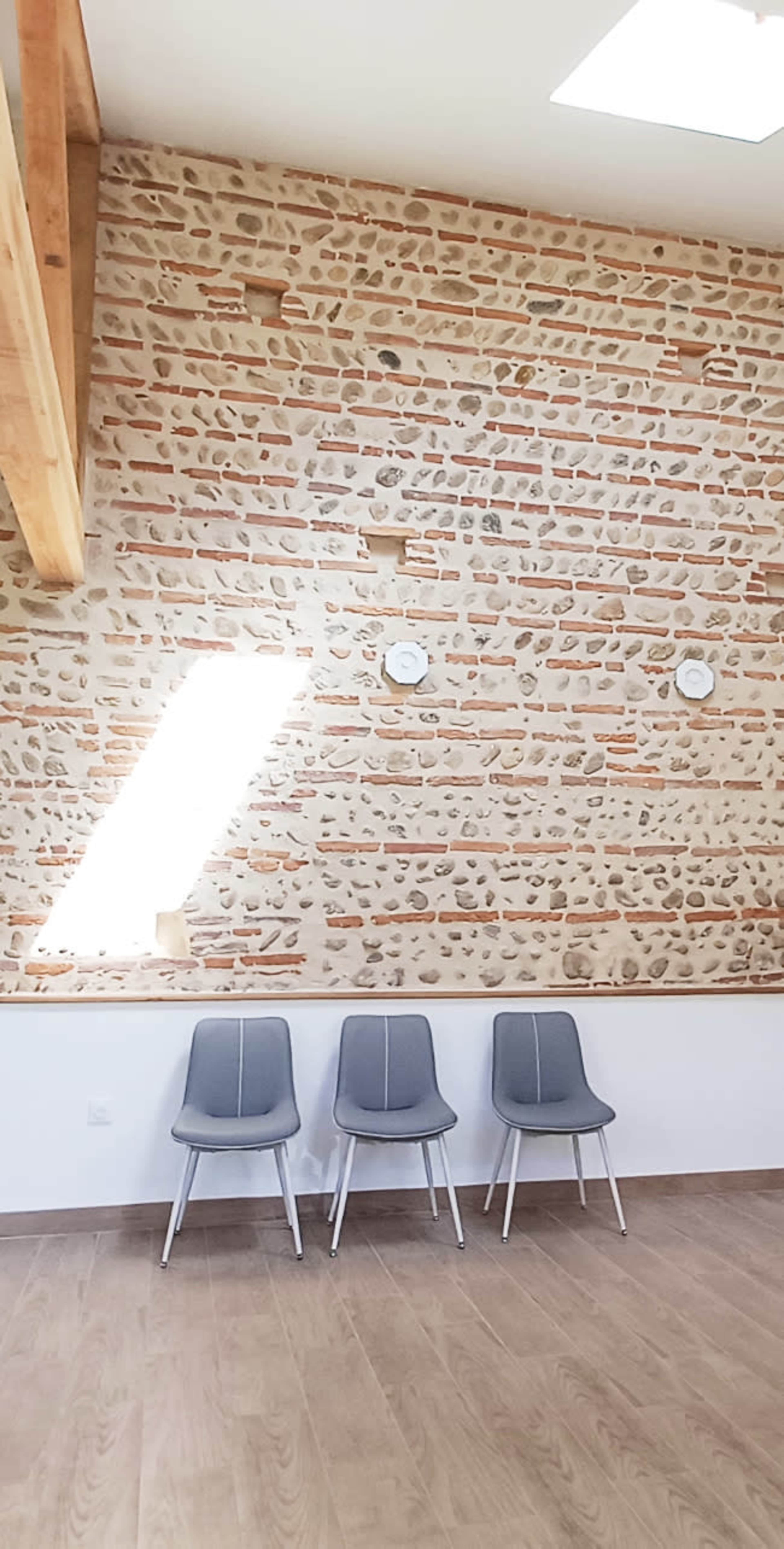 The image shows a room with a textured stone wall, three gray chairs lined up against the wall, and a skylight above.