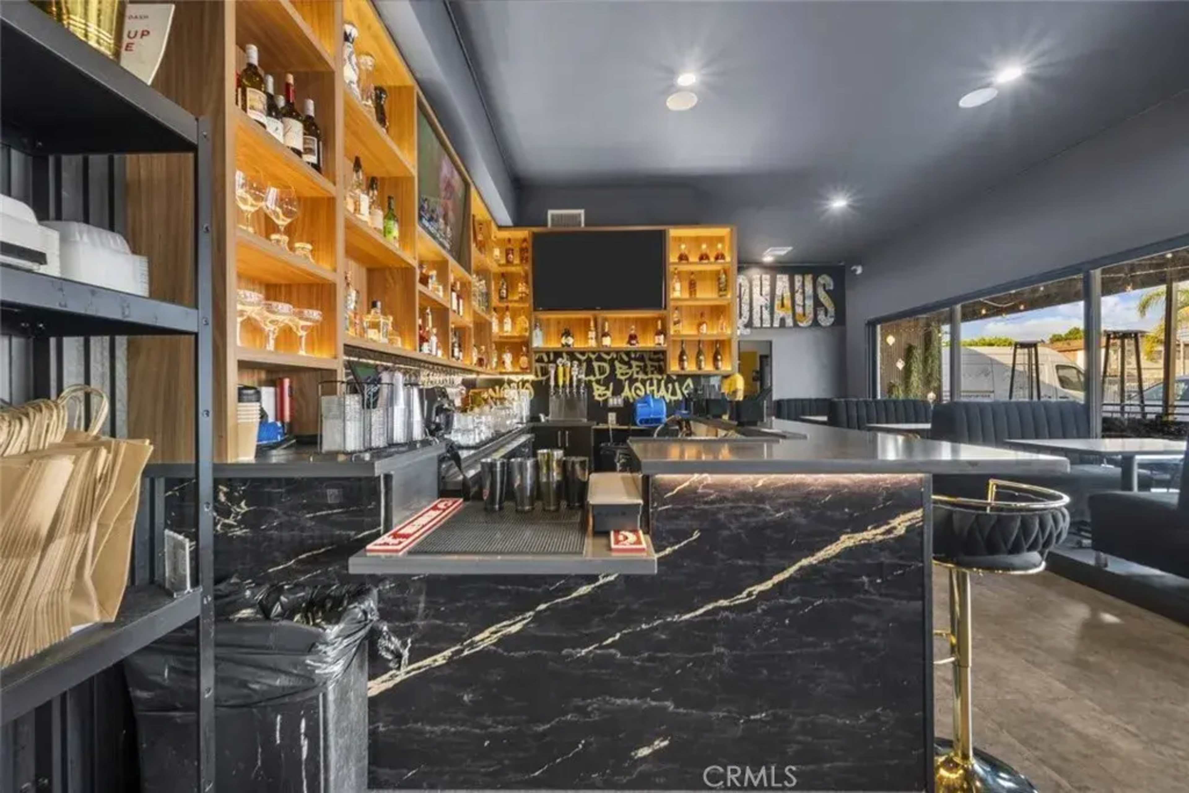 The image shows a modern bar area with a dark marble countertop, wooden shelves displaying various bottles, and a clean, organized layout.