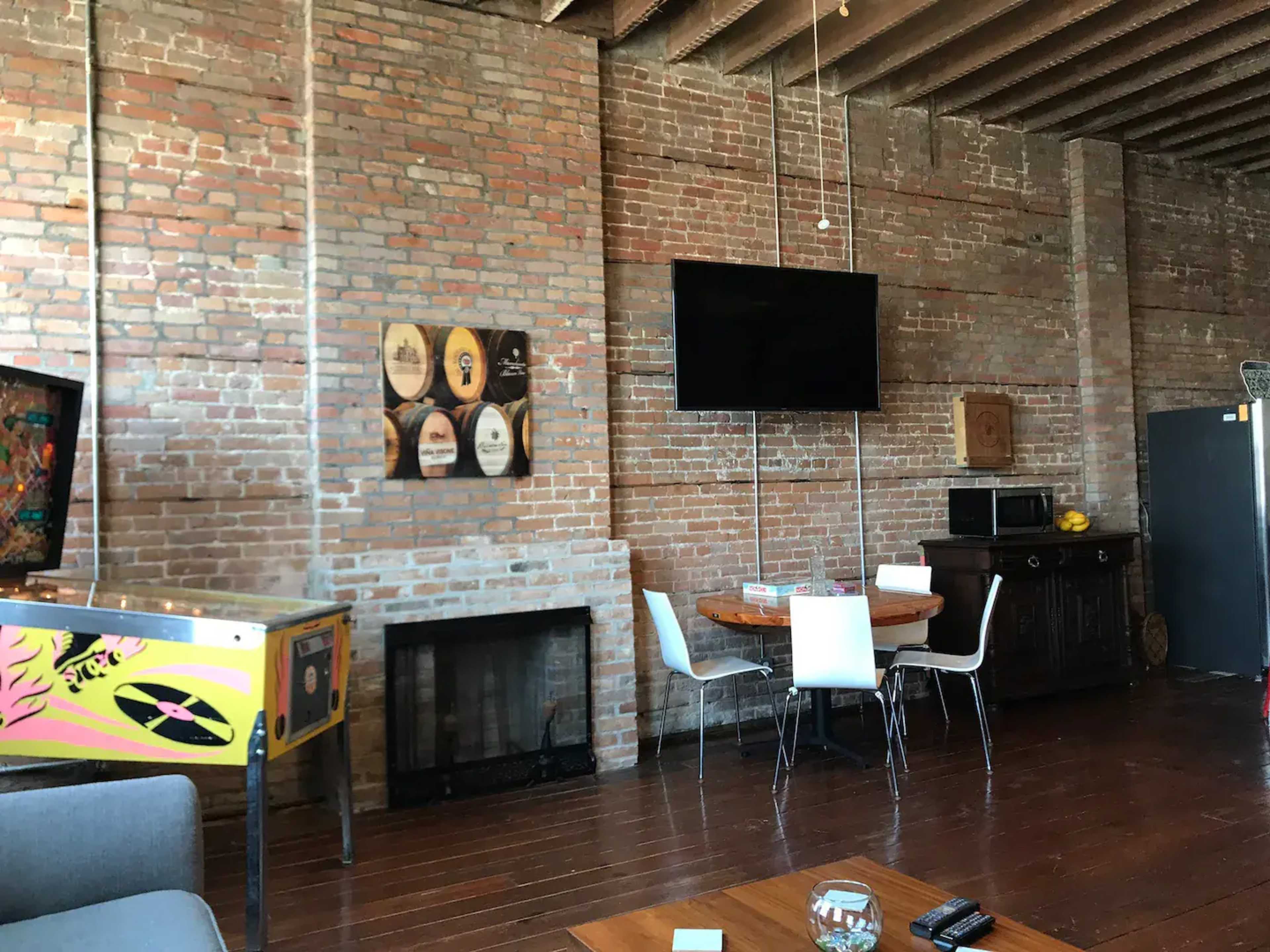 The image shows an interior space with exposed brick walls, a wooden floor, a round dining table with white chairs, a wall-mounted television, and a vintage arcade machine.