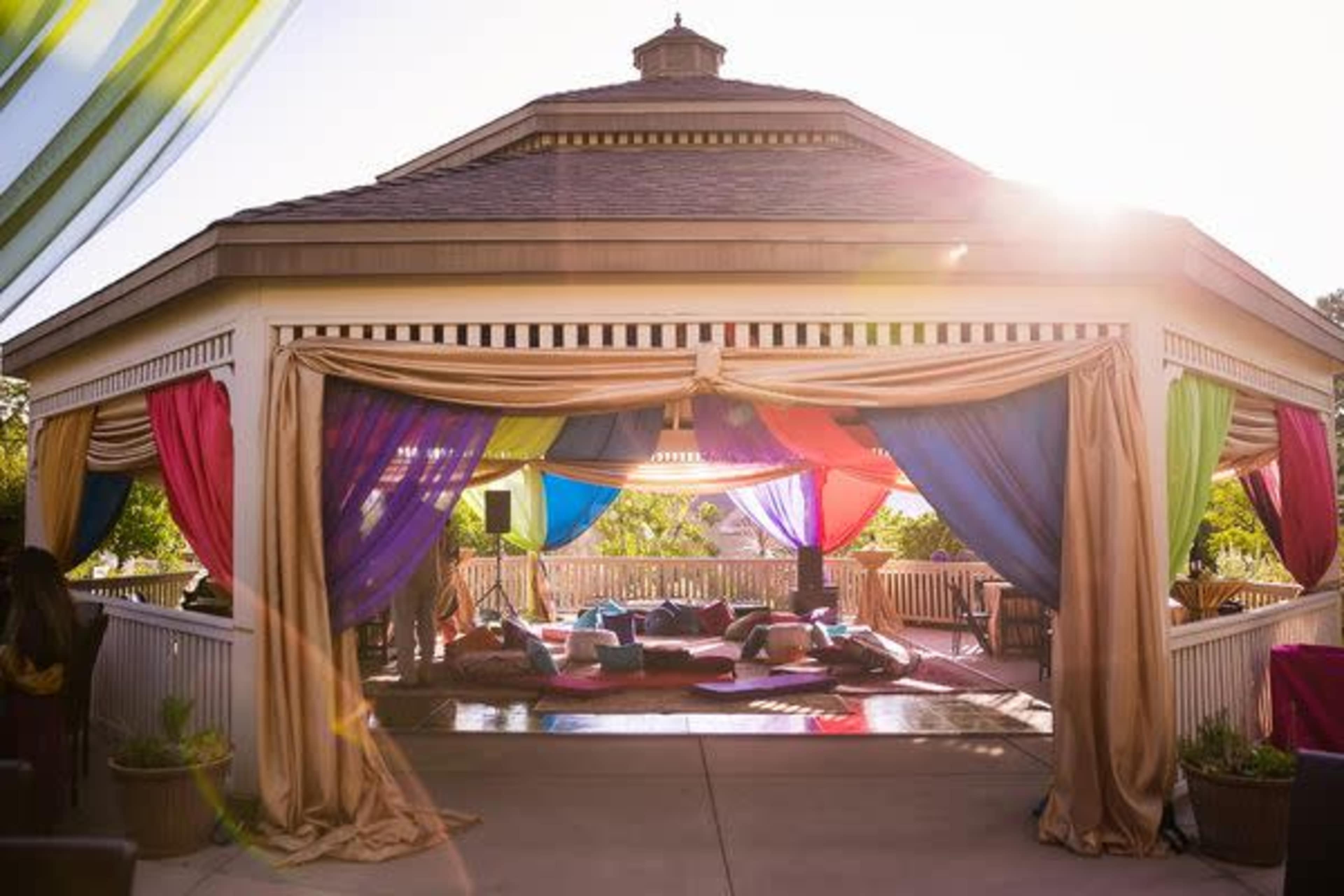 A spacious outdoor pavilion is draped with colorful fabric, creating a vibrant and inviting atmosphere with cushions arranged on the floor.