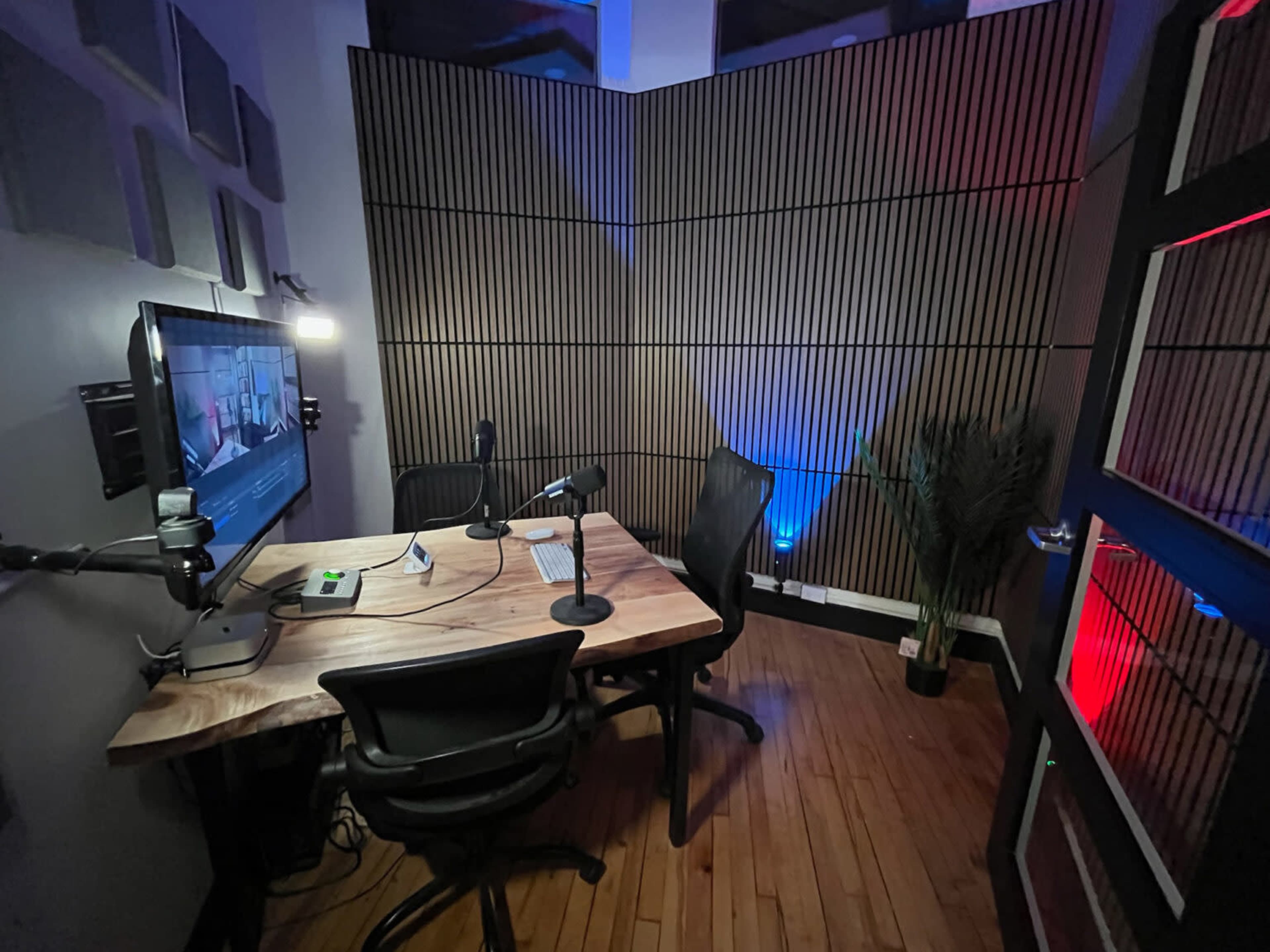 The image shows a small recording studio with a wooden table, two microphones on stands, a monitor displaying content, and ambient lighting in blue and red tones.