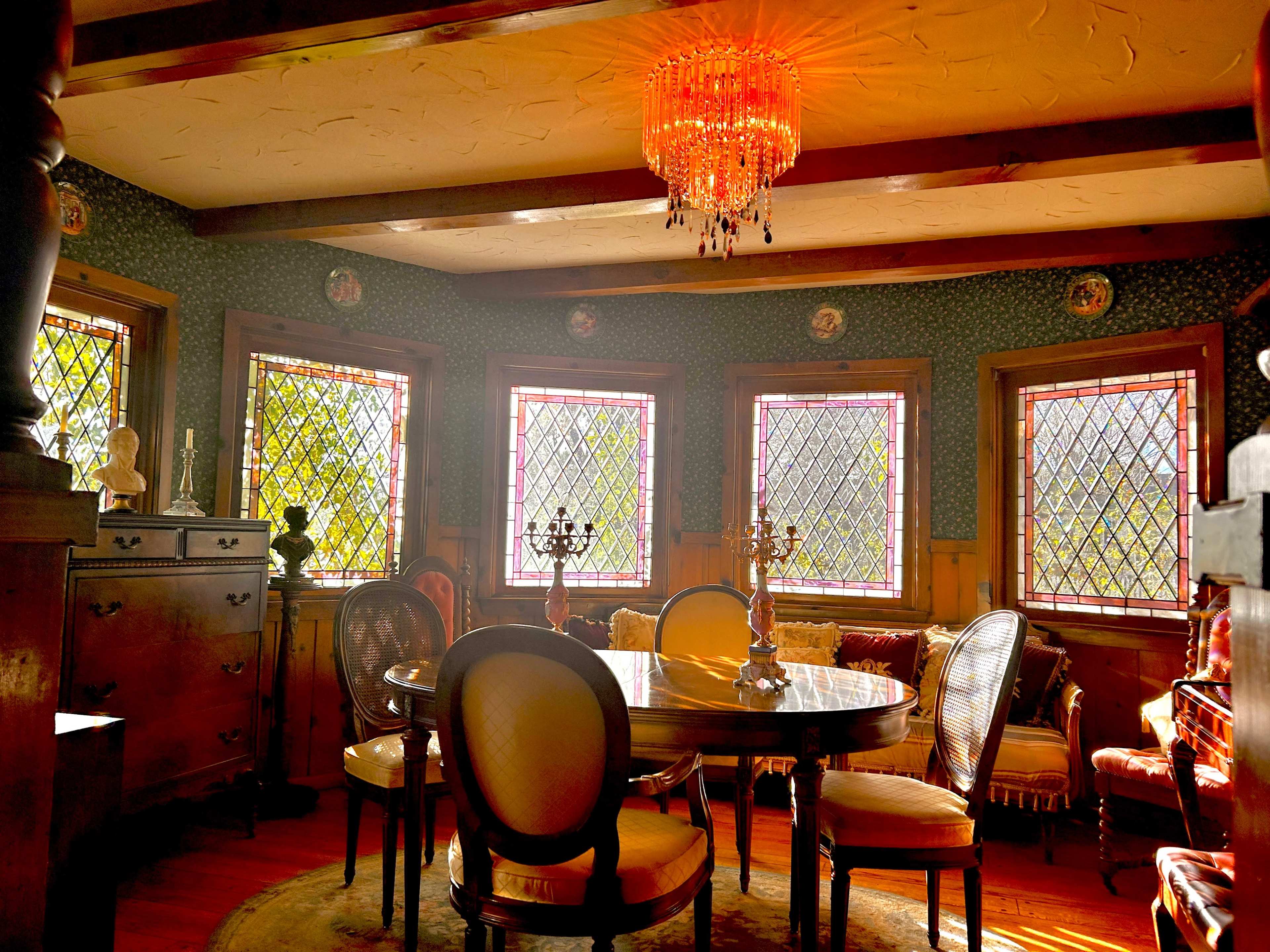 A dining room with a round table surrounded by upholstered chairs, illuminated by a chandelier and featuring large, stained glass windows.