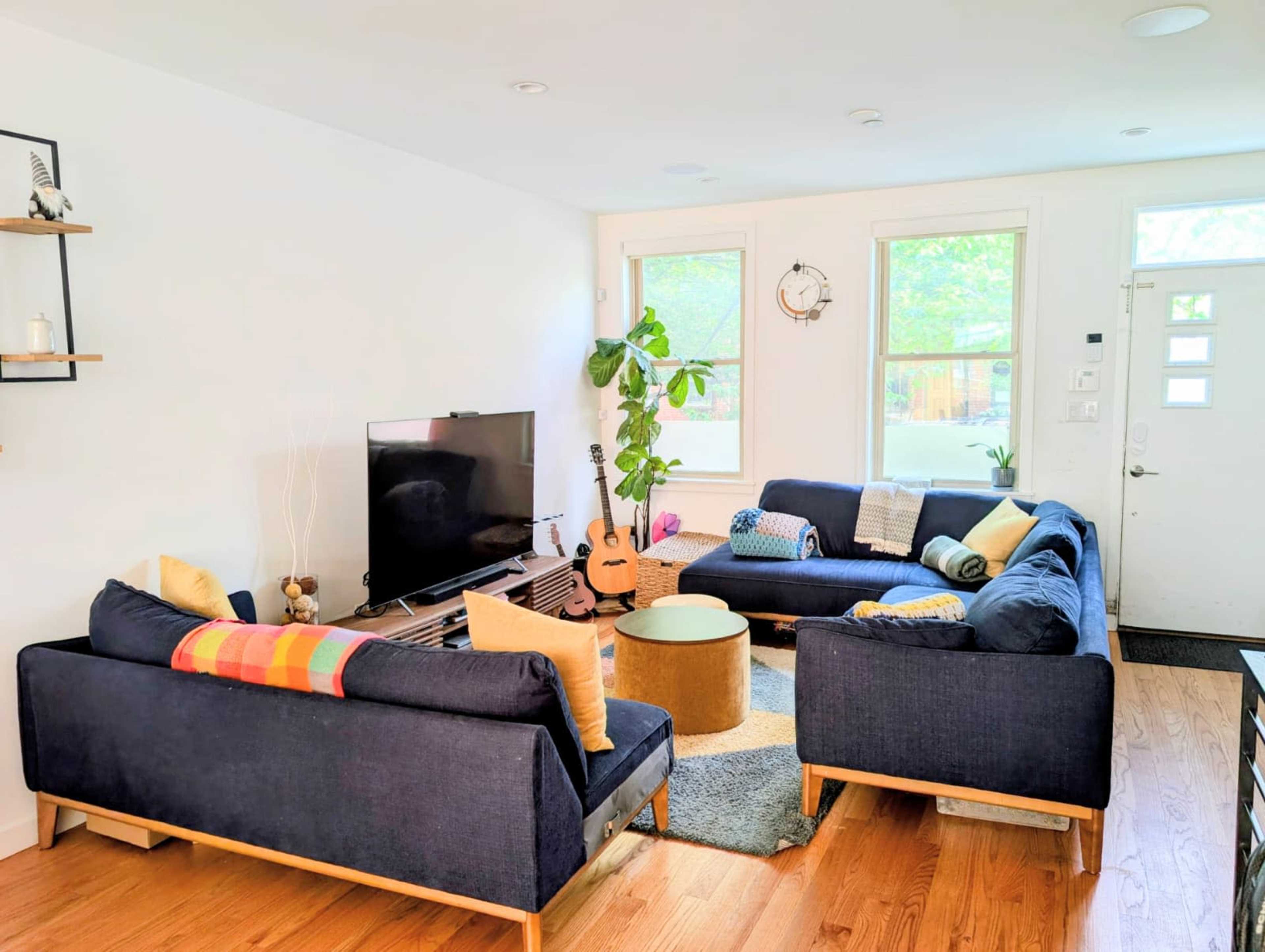 The living room features two navy couches arranged around a central coffee table, with a television on a stand and a guitar nearby, while natural light streams in through the windows.