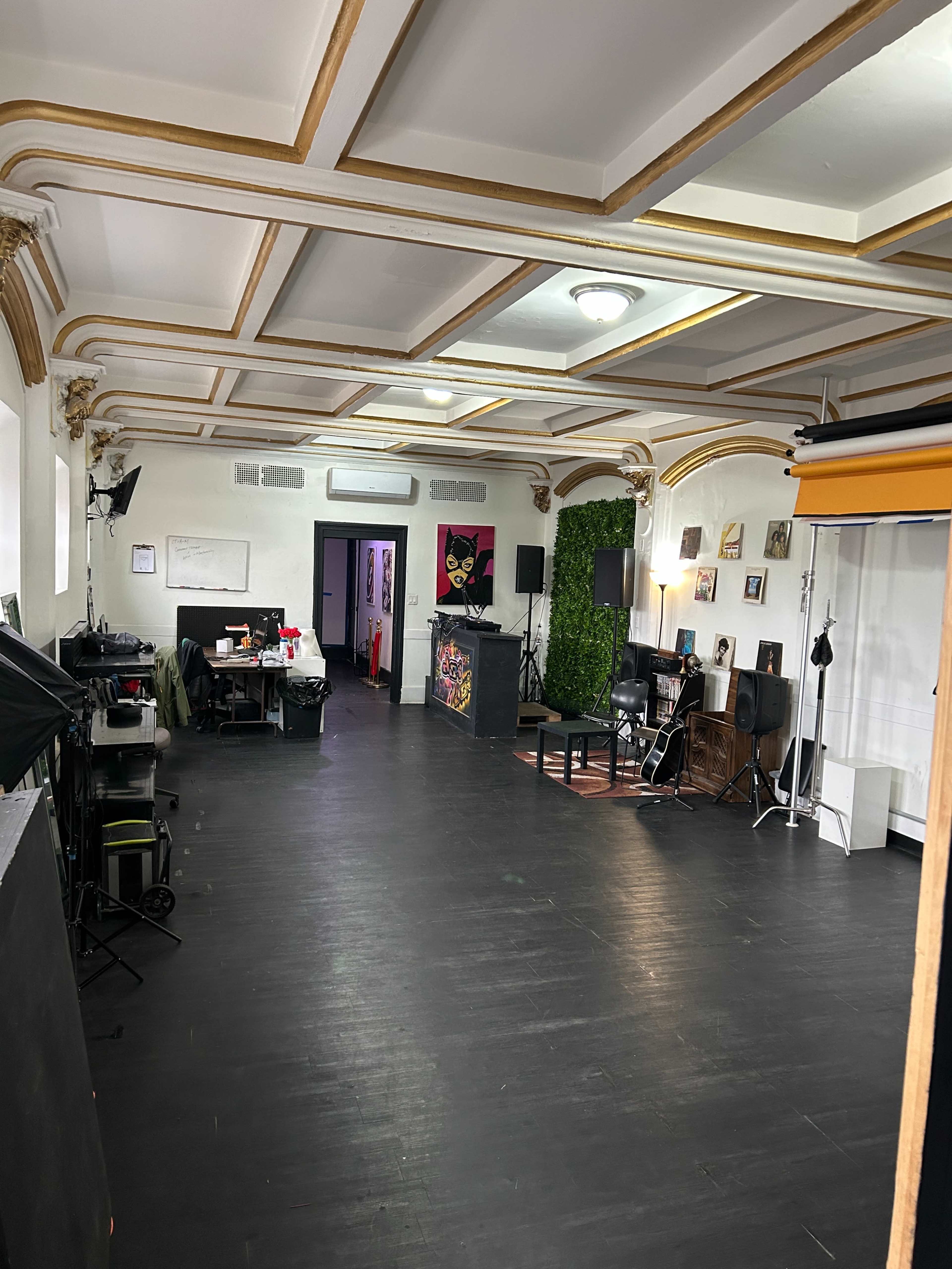 The image shows a spacious interior with dark flooring, featuring various equipment for photography or video production, and a wall adorned with plants and art.
