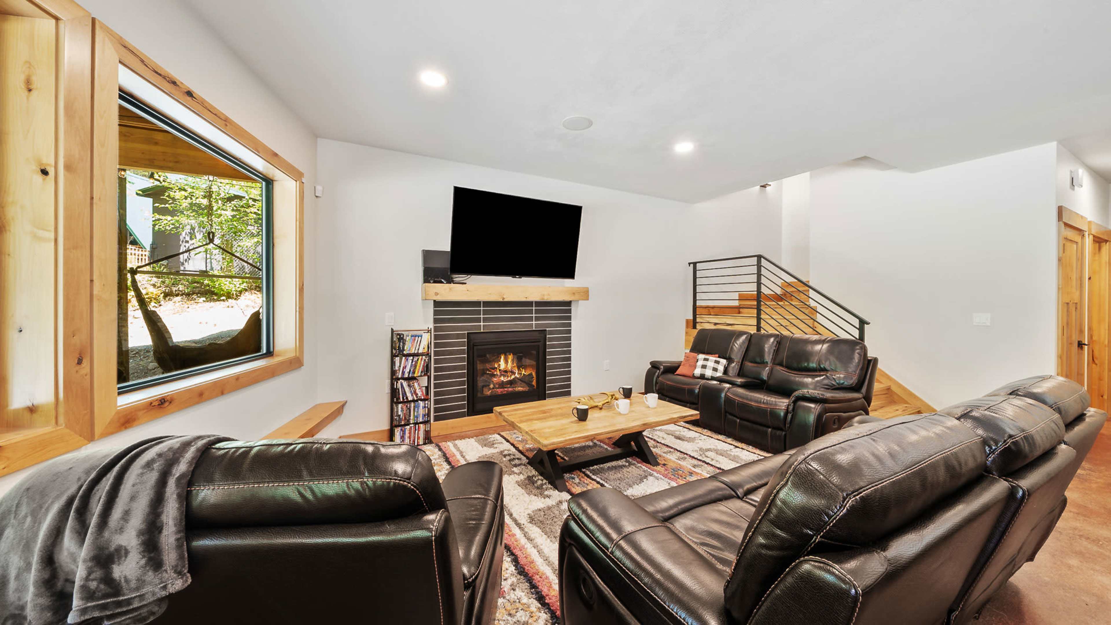 The image shows a modern living room with leather couches, a fireplace, a coffee table, and large windows overlooking greenery.