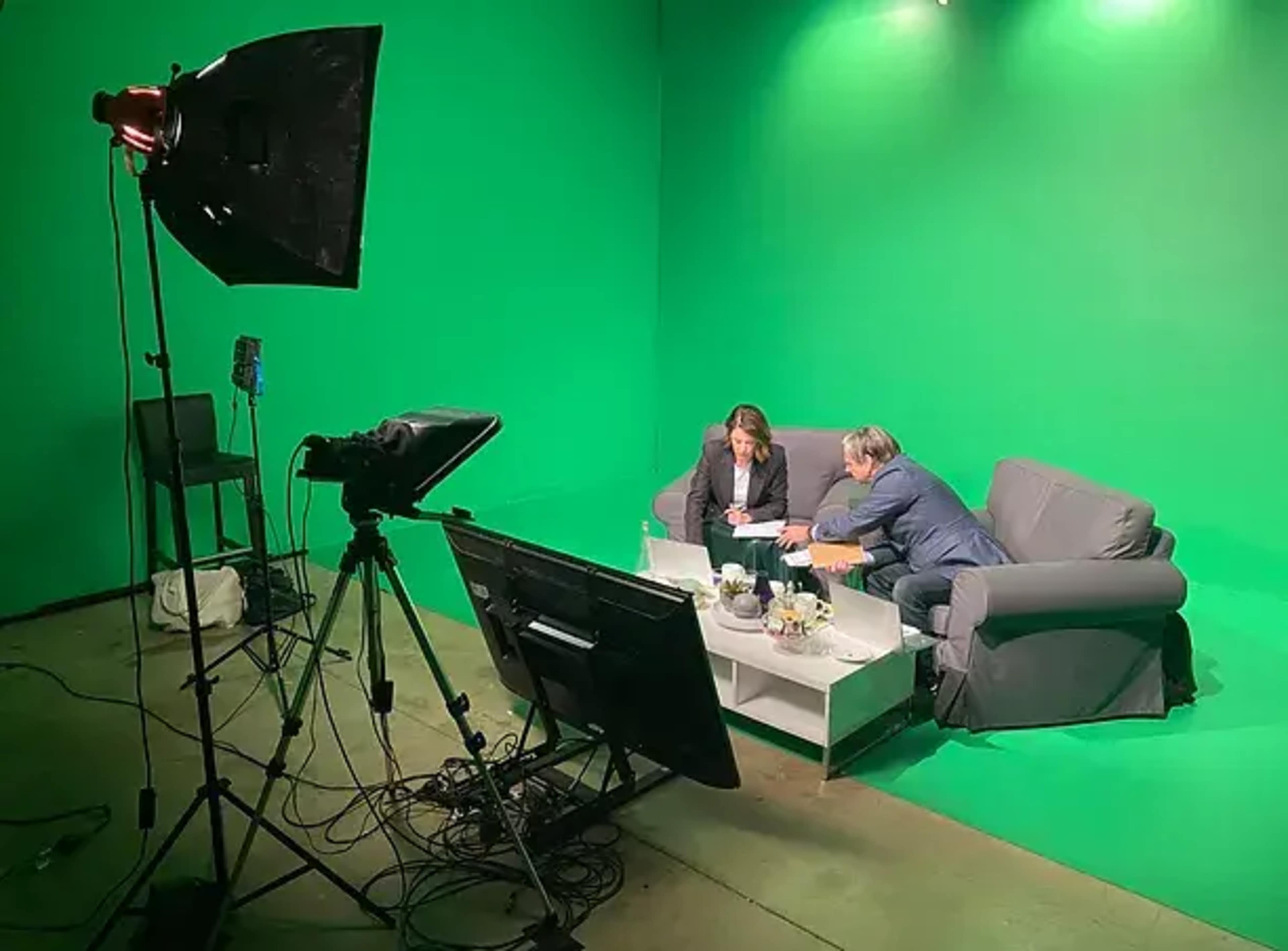 Two people are seated on a gray couch in a green screen studio, discussing something over a laptop on a coffee table.