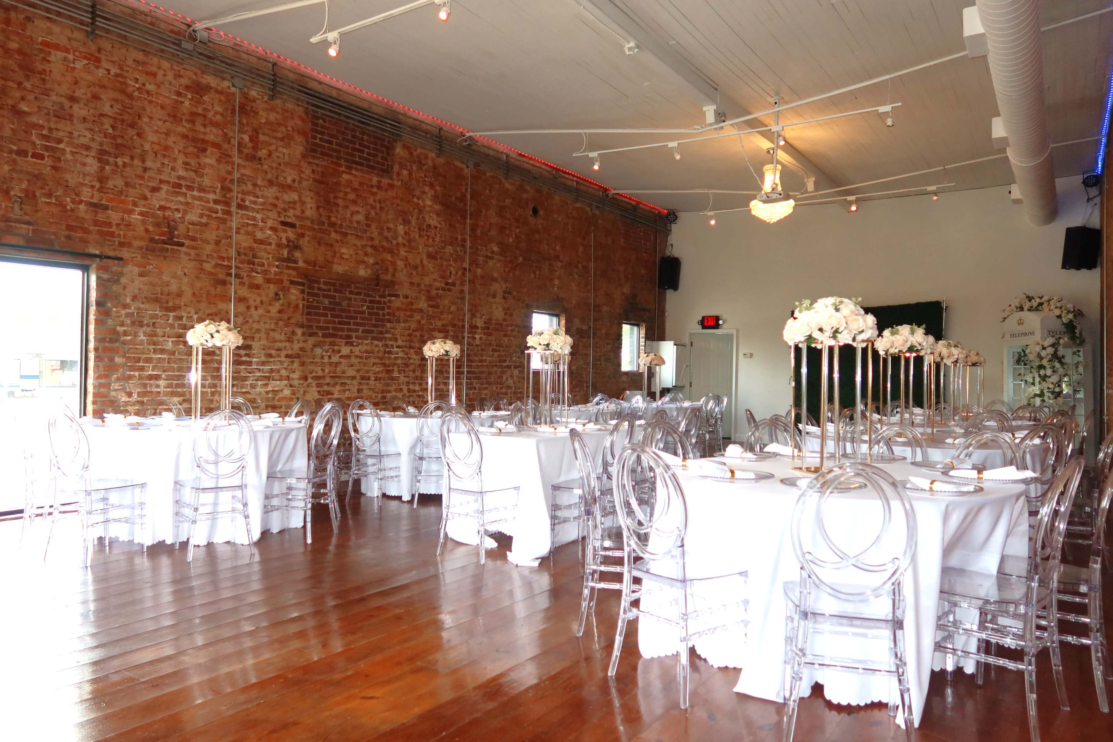 A spacious indoor venue features round tables with white tablecloths and clear acrylic chairs, adorned with floral centerpieces against a backdrop of exposed brick walls.