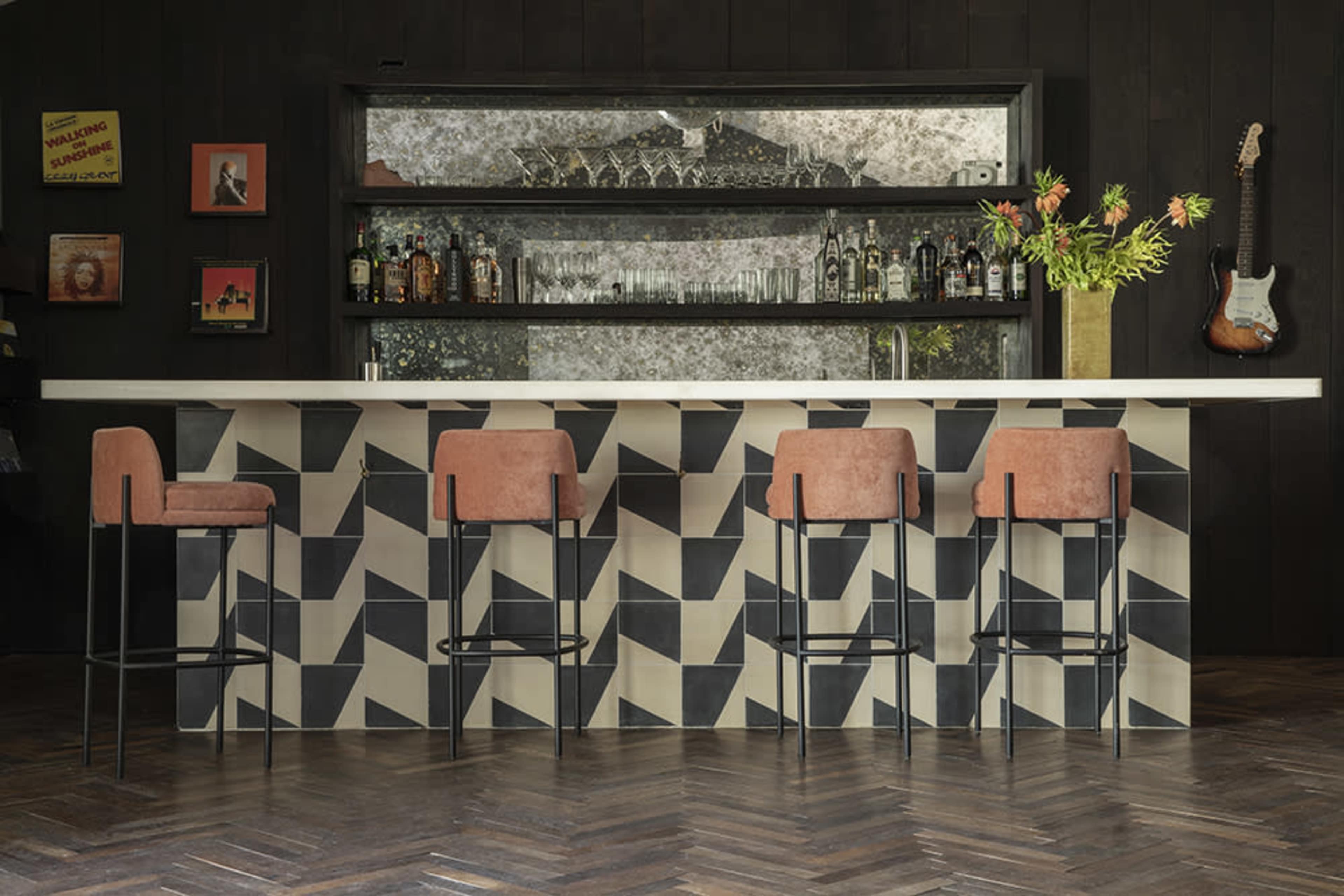 A modern bar with a geometric-patterned countertop, four upholstered stools, and a mirrored back display containing various bottles.
