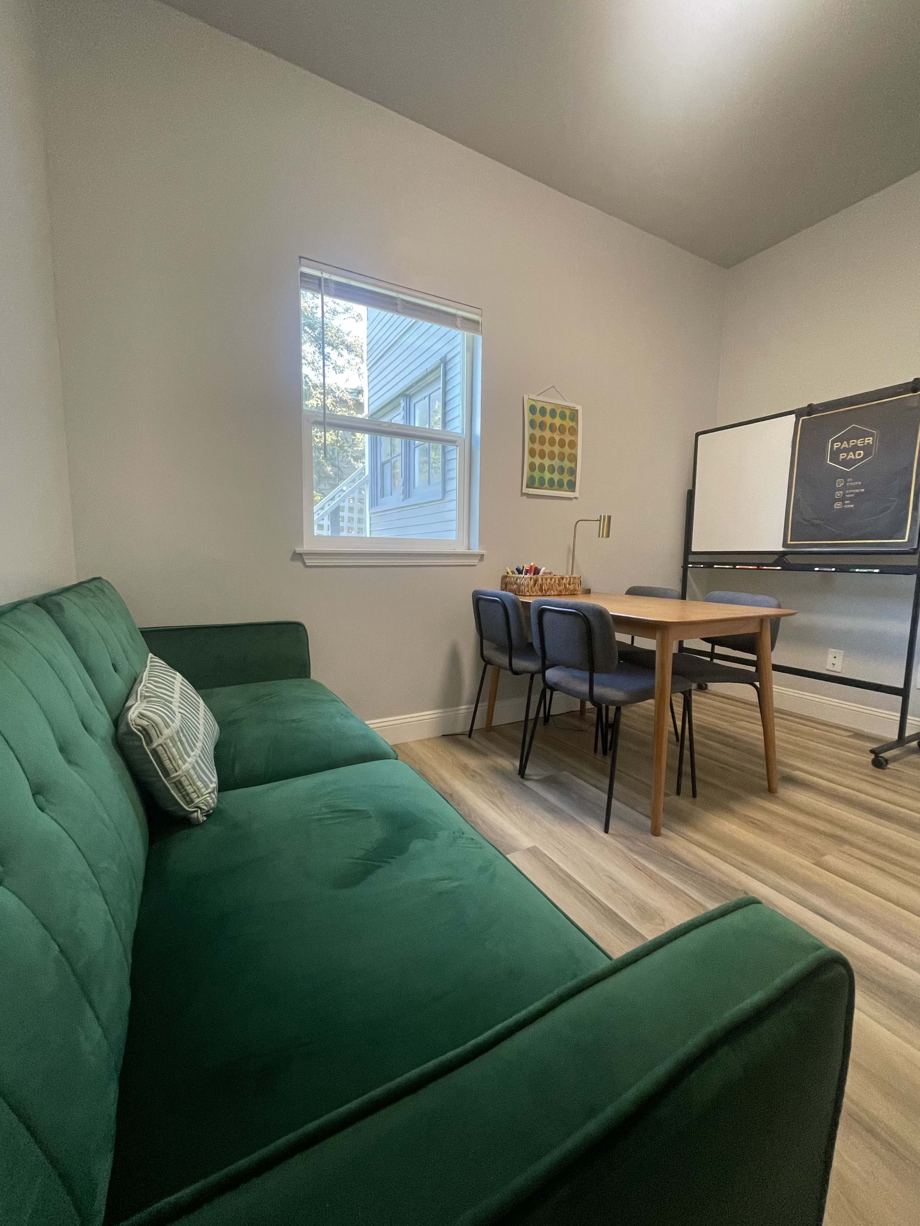 A small room features a green sofa, a wooden table with chairs, and a window with natural light.