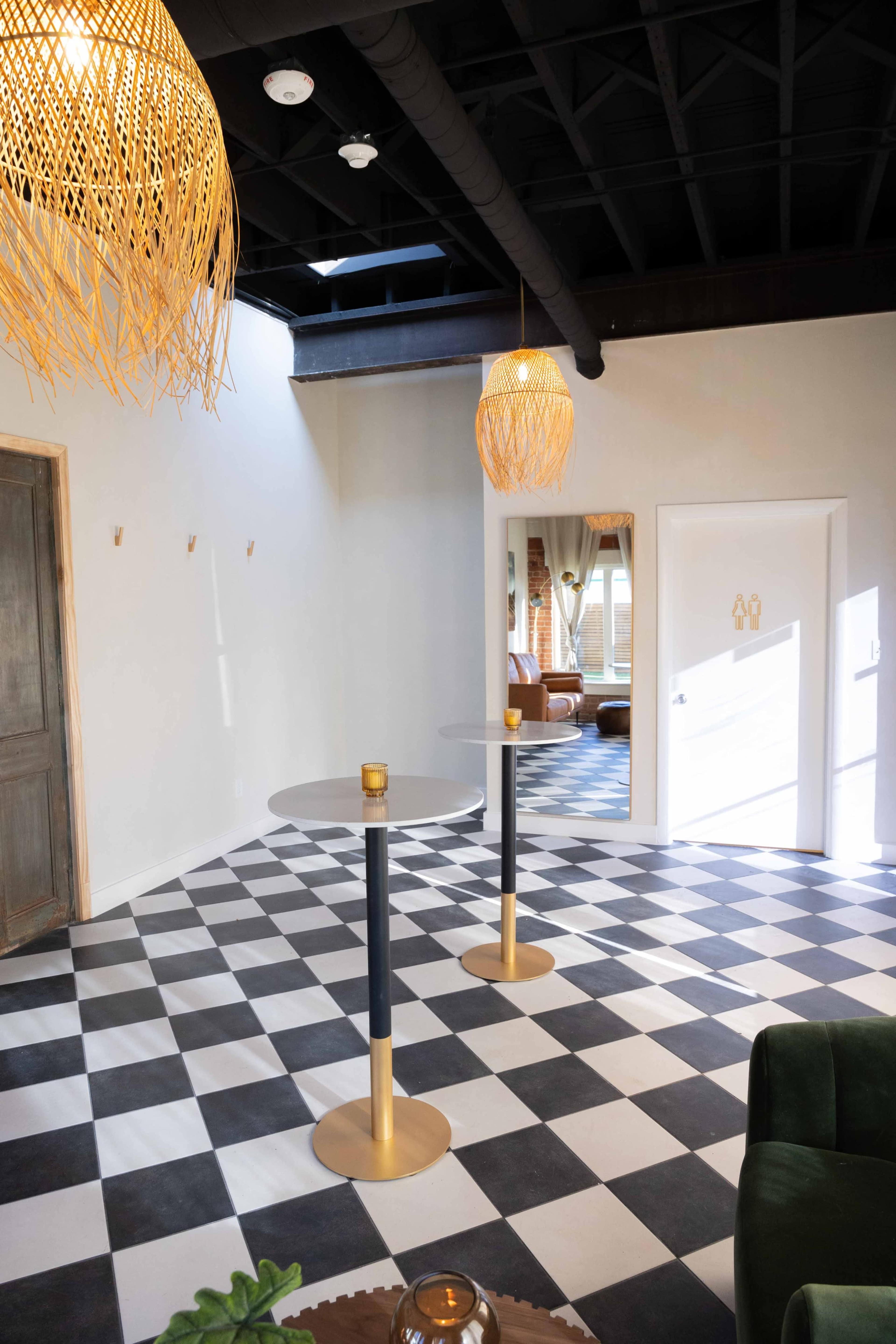 The image shows a room with checkered black and white flooring, two gold-accented standing tables, and pendant lights hanging from the ceiling.