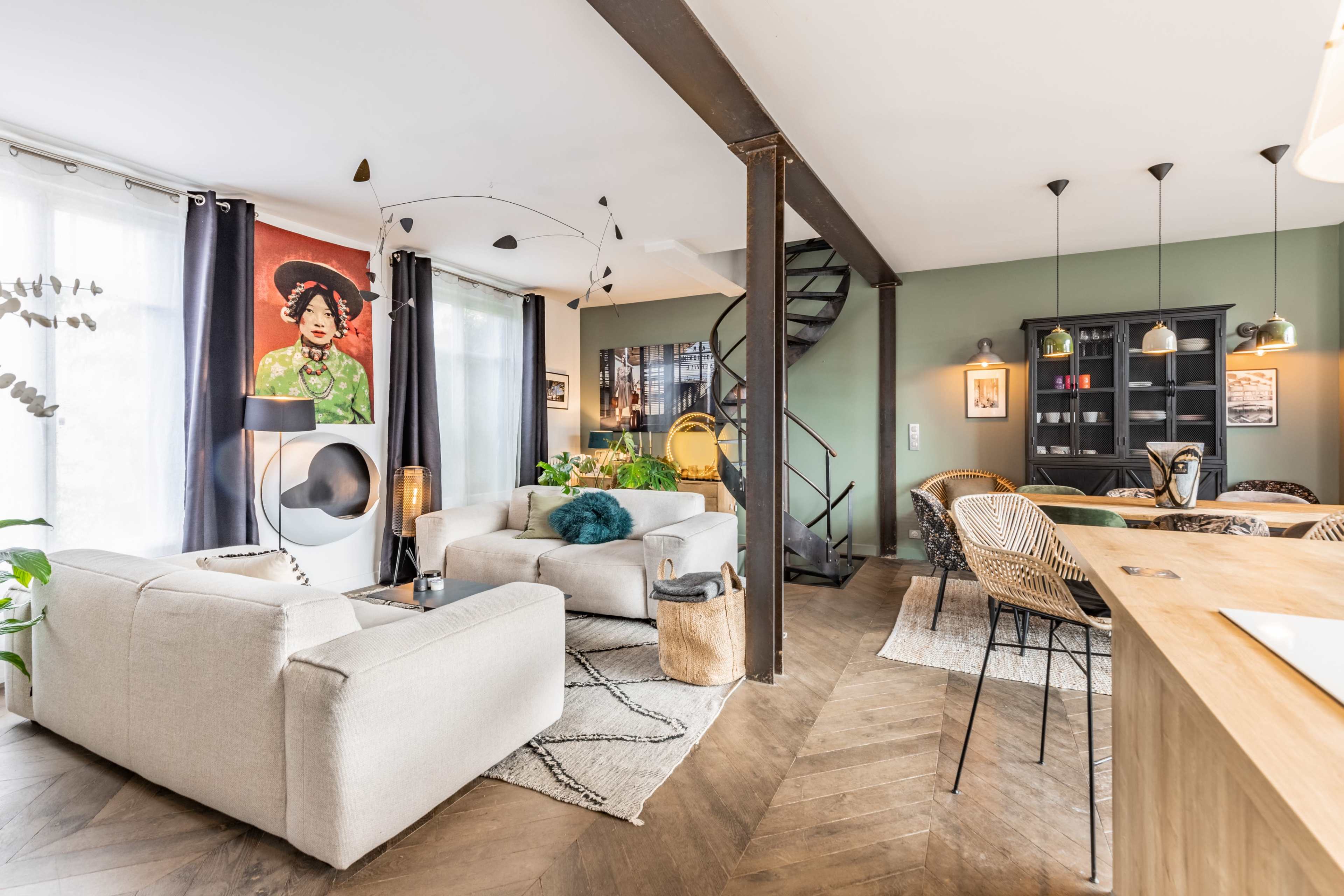 A modern living space featuring two light-colored sofas, a spiral staircase, and a dining area with wicker chairs, decorated with a large colorful wall art and greenery.