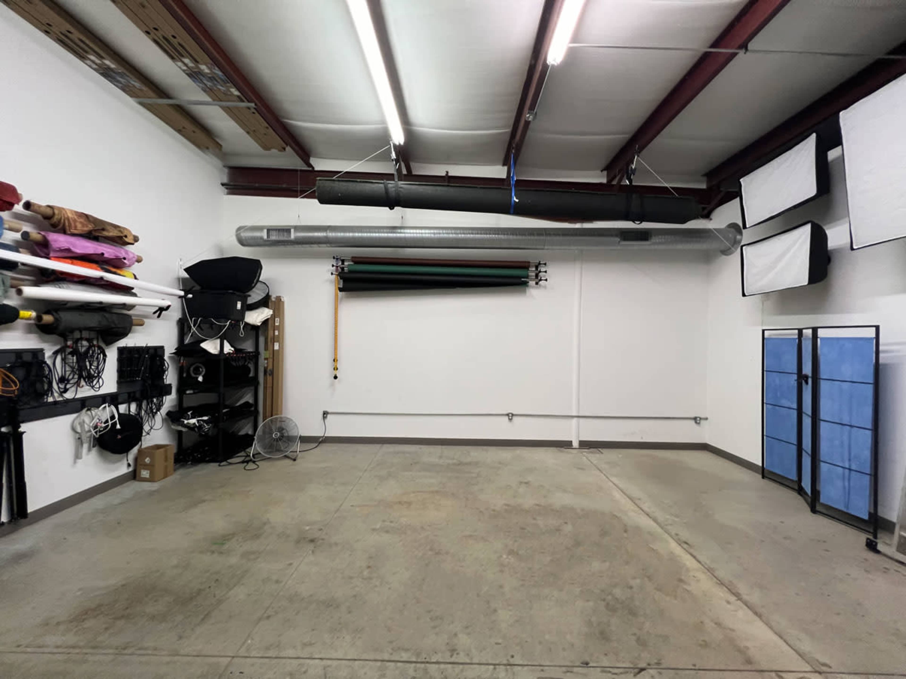 The image shows a spacious studio with white walls, concrete flooring, and various backdrop rolls and equipment organized along the walls.