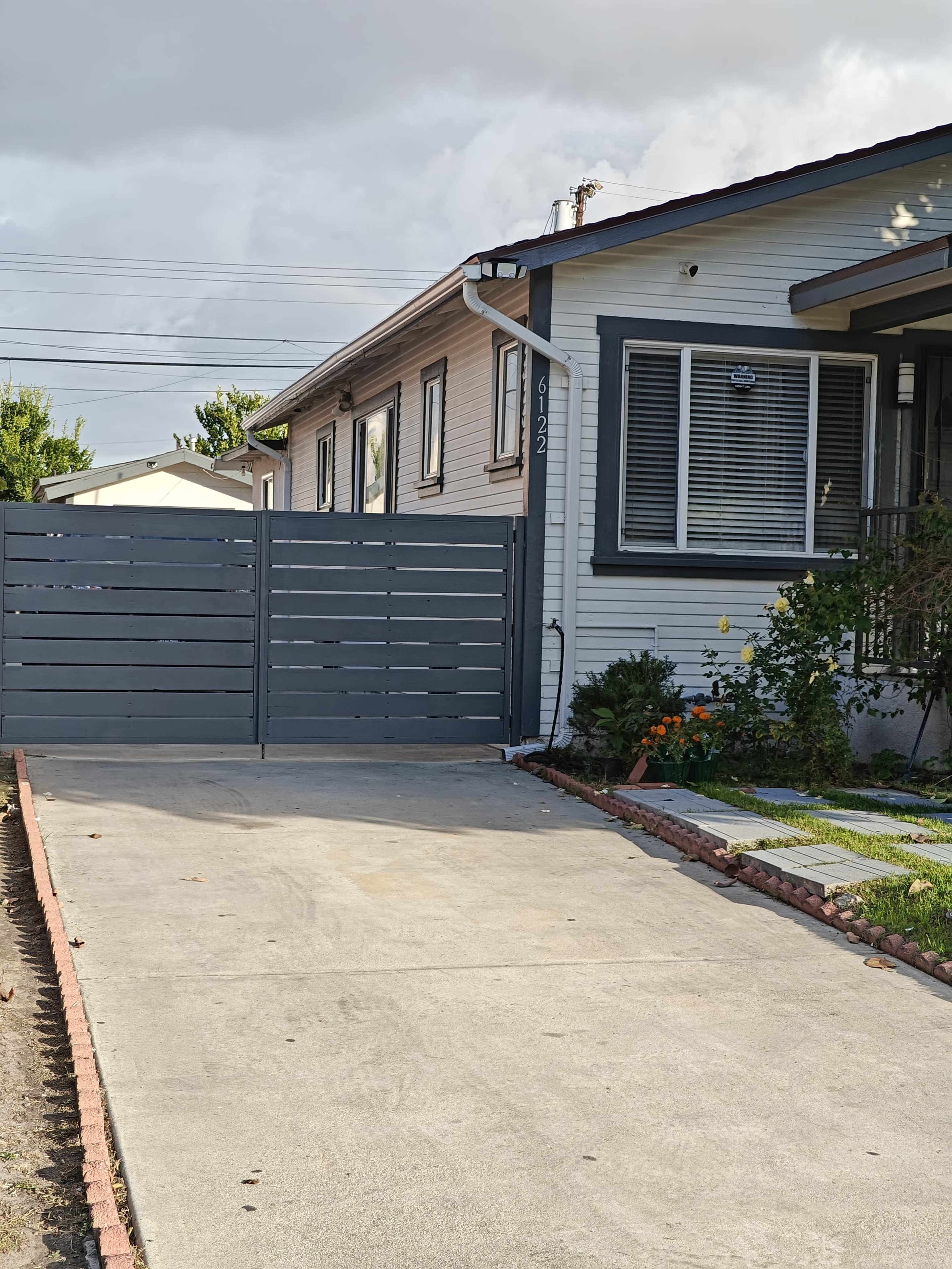 A gray wooden gate is closed at the entrance of a driveway leading to a single-story house with a well-maintained front yard and flower beds.