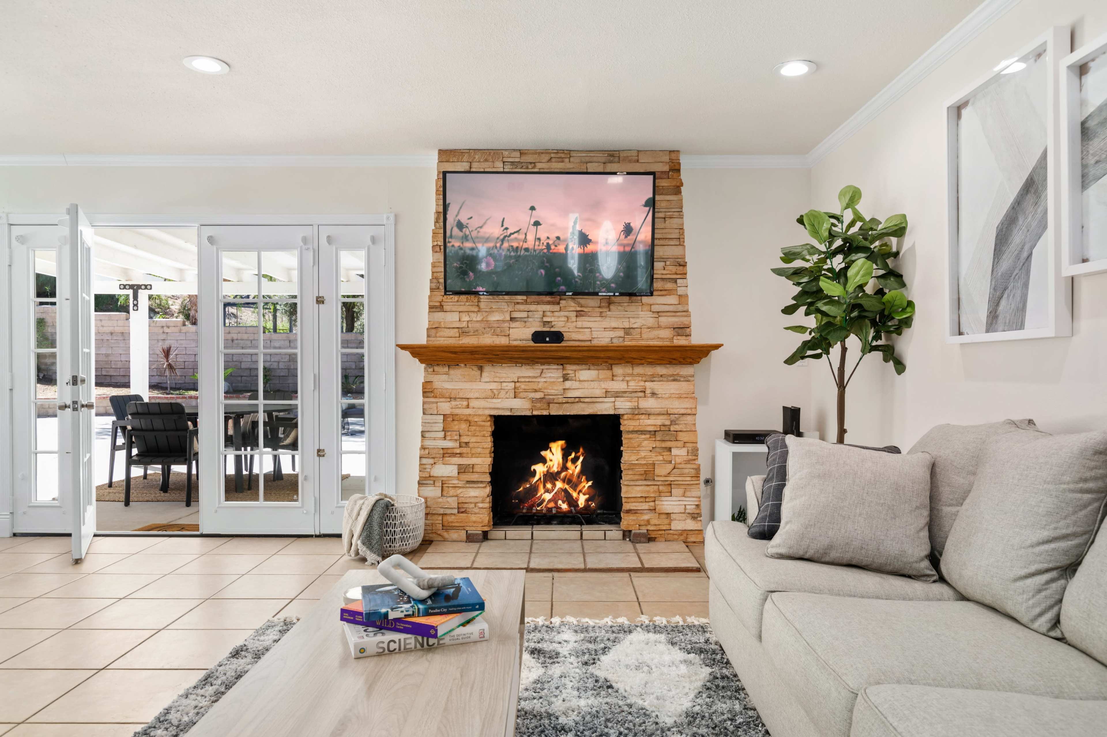 A living room features a stone fireplace with a TV mounted above it, a gray couch, and a large window with doors leading to an outdoor seating area.