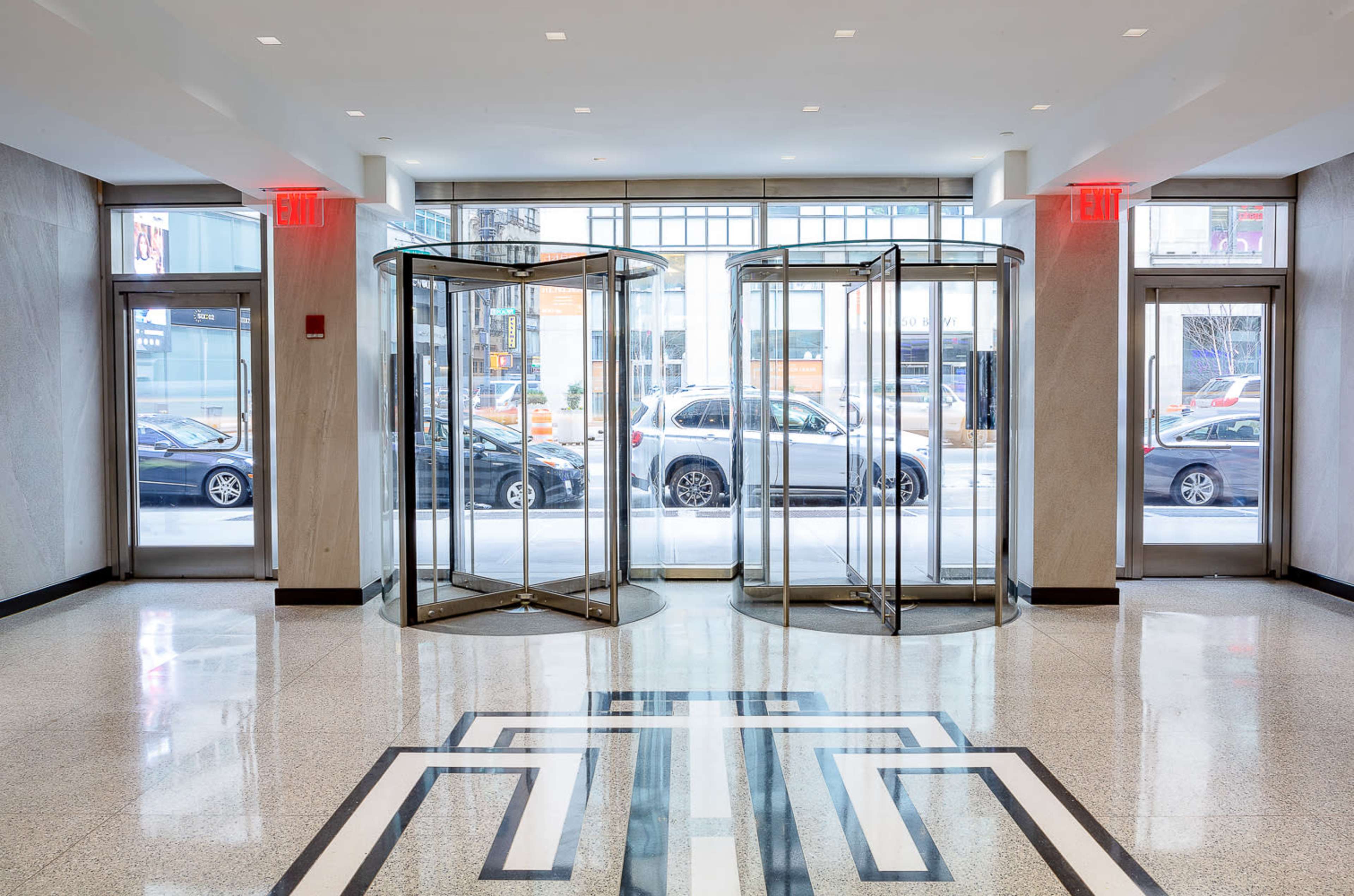The image shows a bright, spacious lobby with three revolving doors and a patterned floor design, flanked by red emergency exit lights.