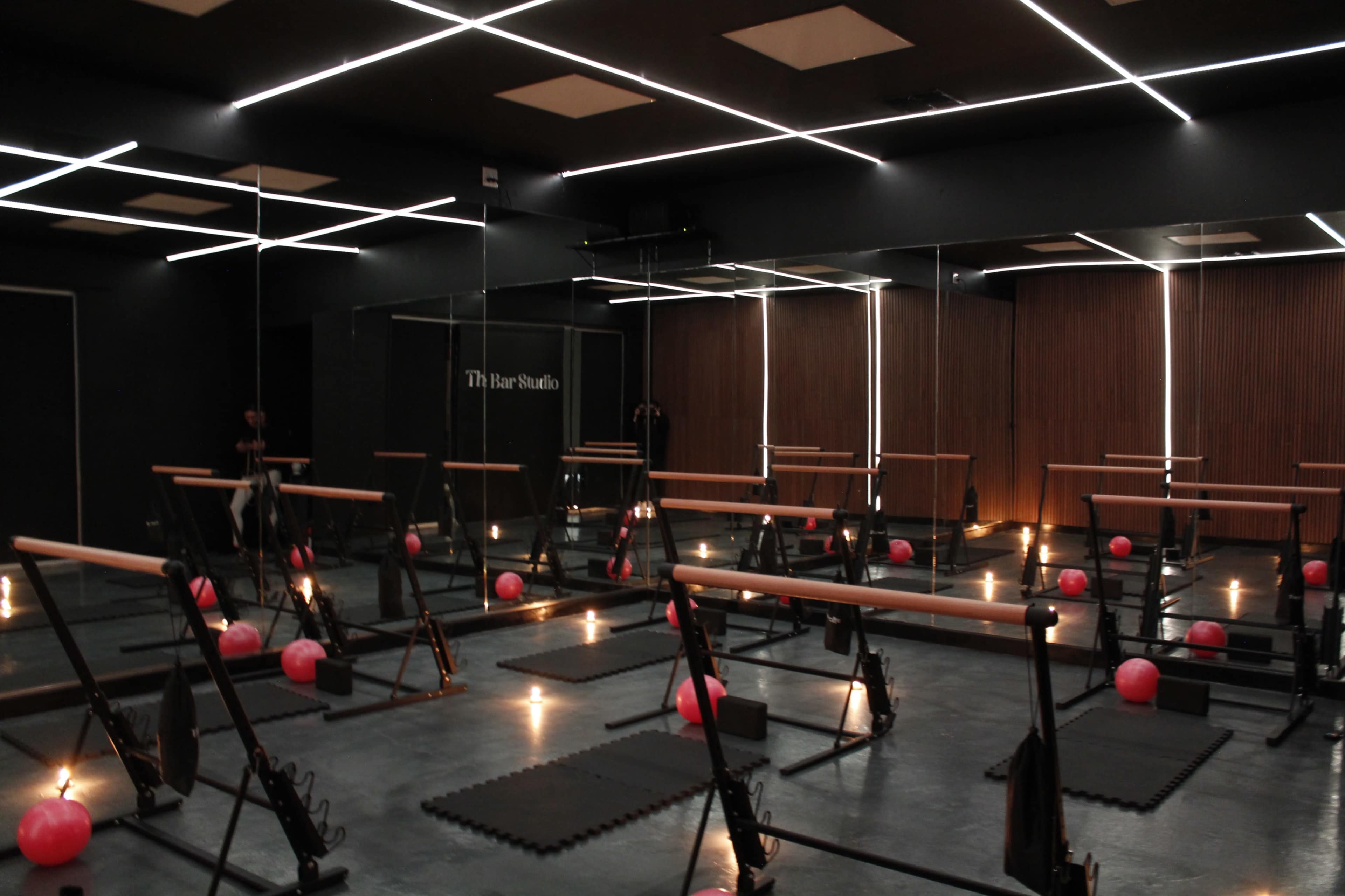 A dimly lit exercise studio with mirrored walls, wooden accents, and various fitness equipment arranged neatly, including pink exercise balls and resistance bands.