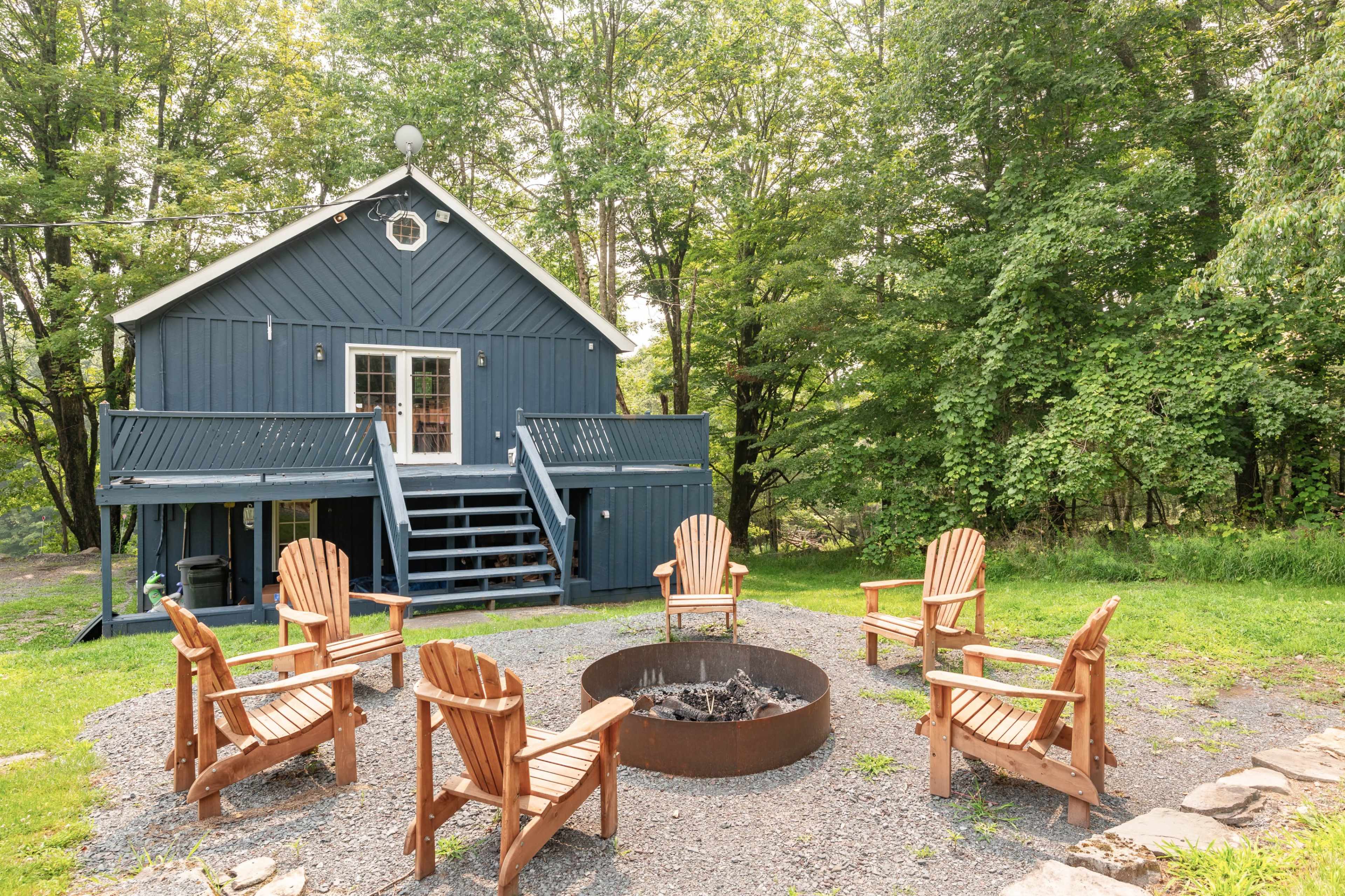 A blue cabin with a front porch is surrounded by a stone fire pit and several wooden chairs in a wooded area.