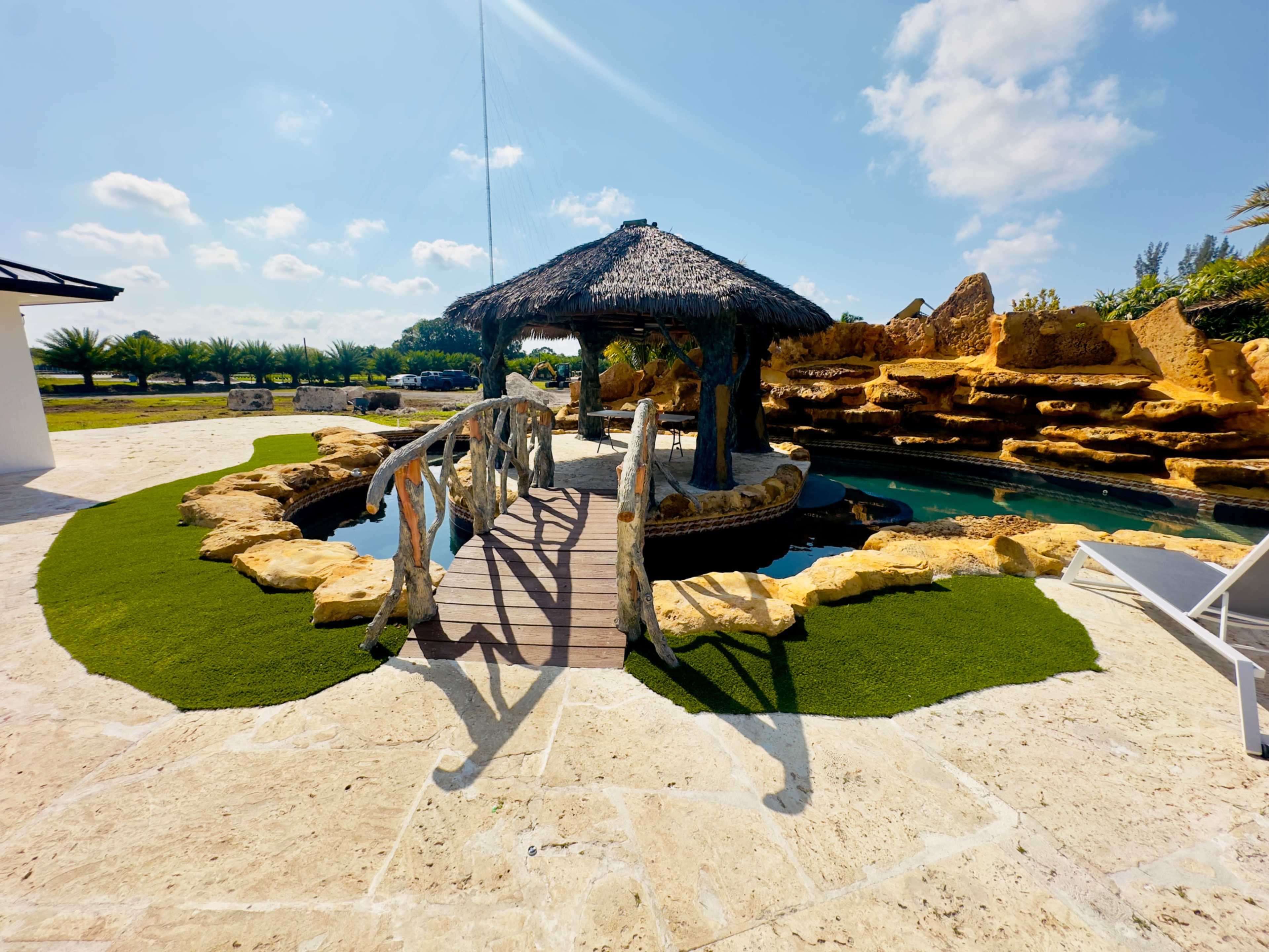 The image shows a thatched gazebo surrounded by artificial grass and rocky landscaping near a swimming pool.
