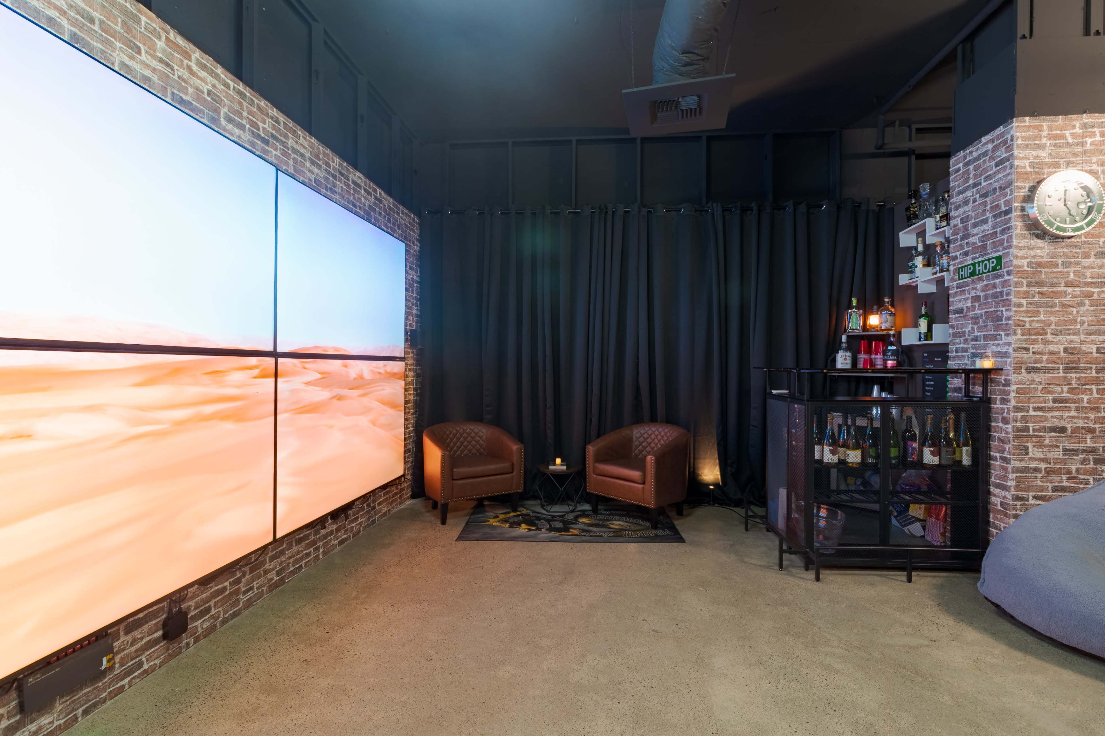 The image shows a modern lounge space featuring two brown leather chairs, a large wall-mounted screen displaying a desert landscape, and a corner bar with various bottles.
