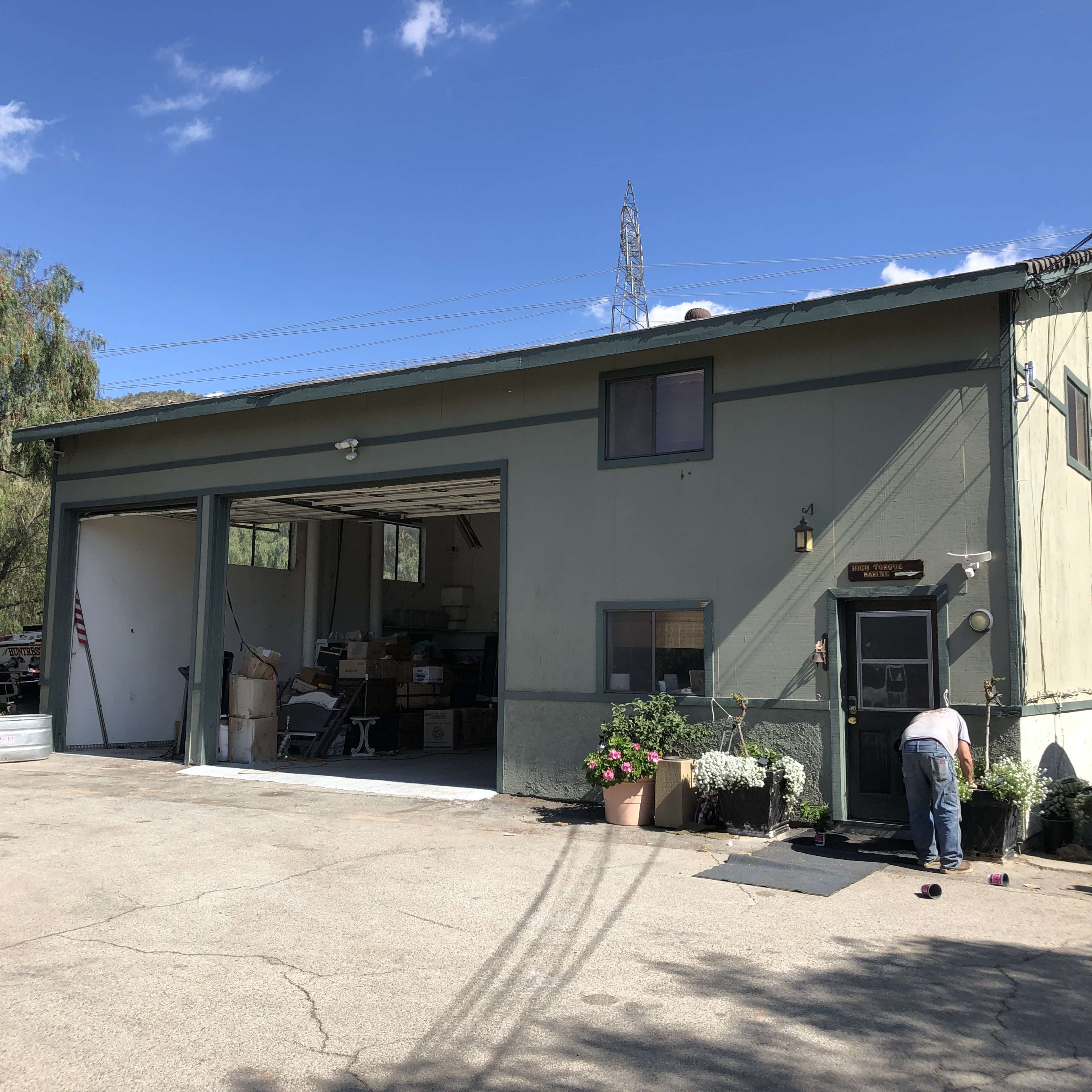 A man is tending to potted plants in front of a large, light green garage building with a partially open door.