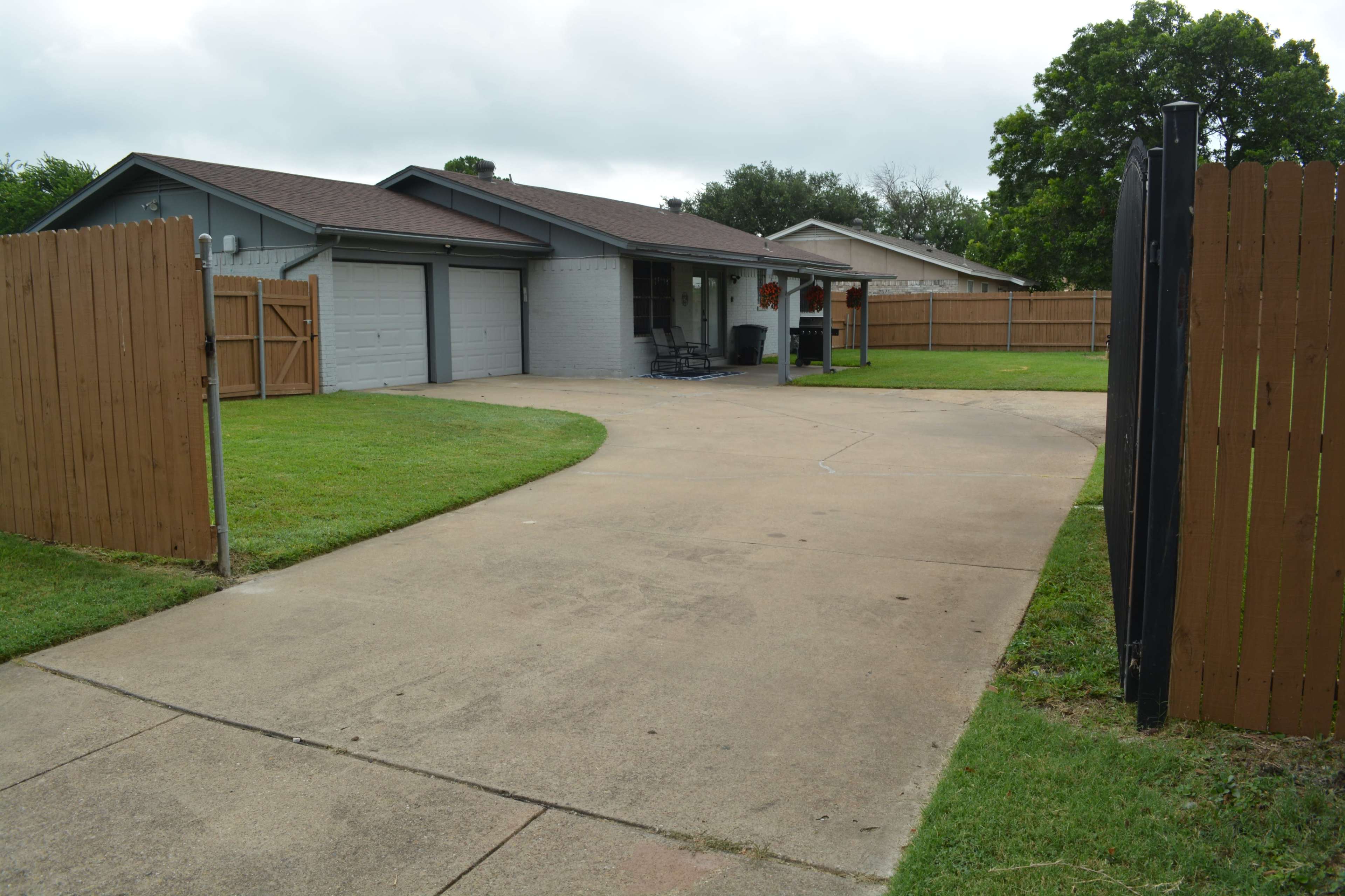 A single-story house with a two-car garage is set back from a concrete driveway, surrounded by a grassy yard and fenced on both sides.