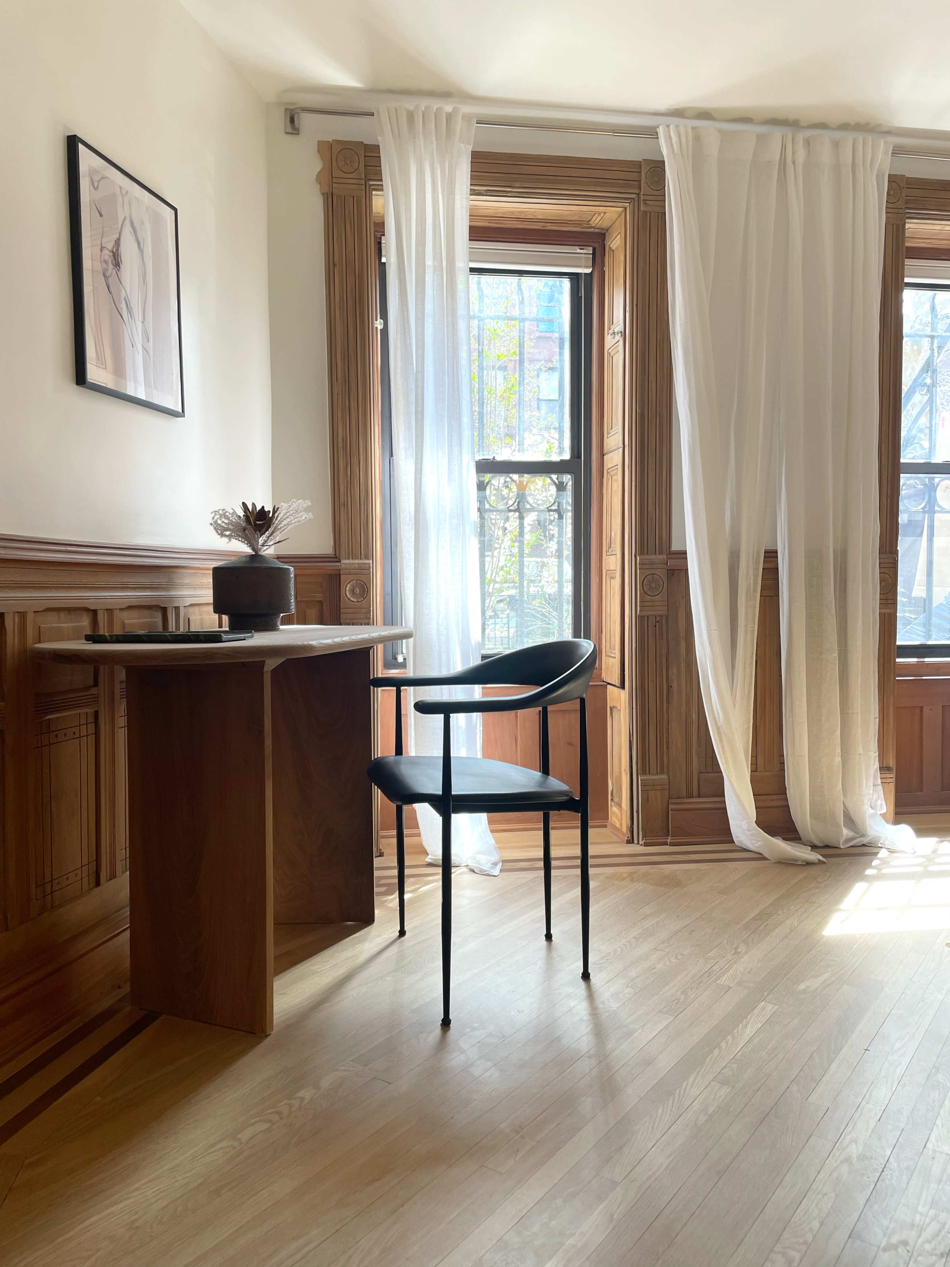 A wooden table and a black chair are placed beside a large window with sheer curtains in a well-lit room with wooden trim.