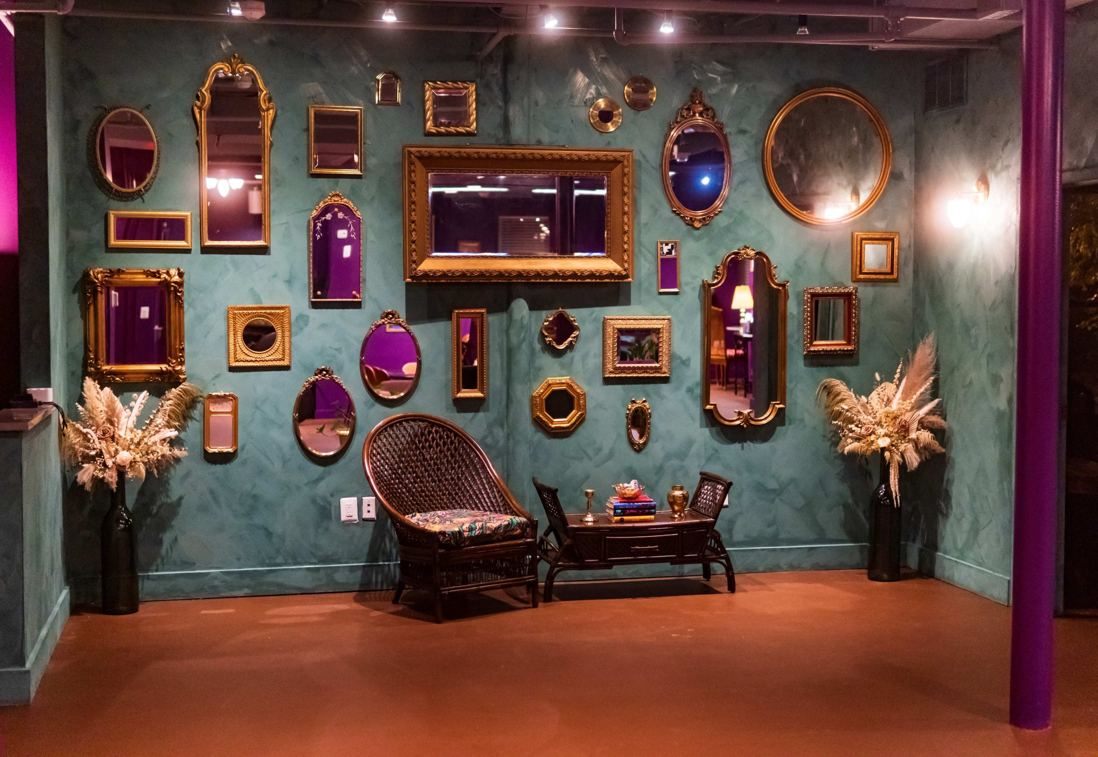 A wall adorned with various decorative mirrors and framed artwork, accompanied by a wicker chair and potted plants.