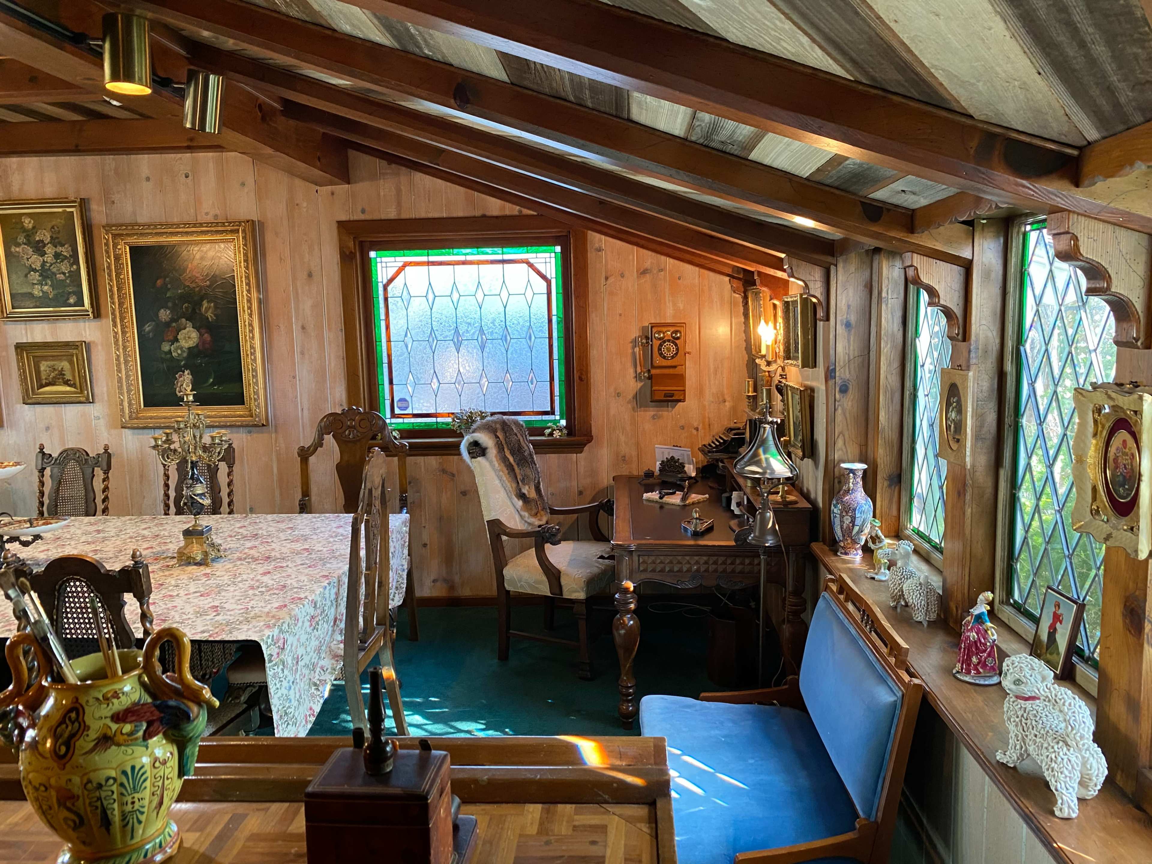 A rustic room with wood-paneled walls, a table covered with a floral tablecloth, vintage furnishings, and decorative items displayed on shelves and a desk.