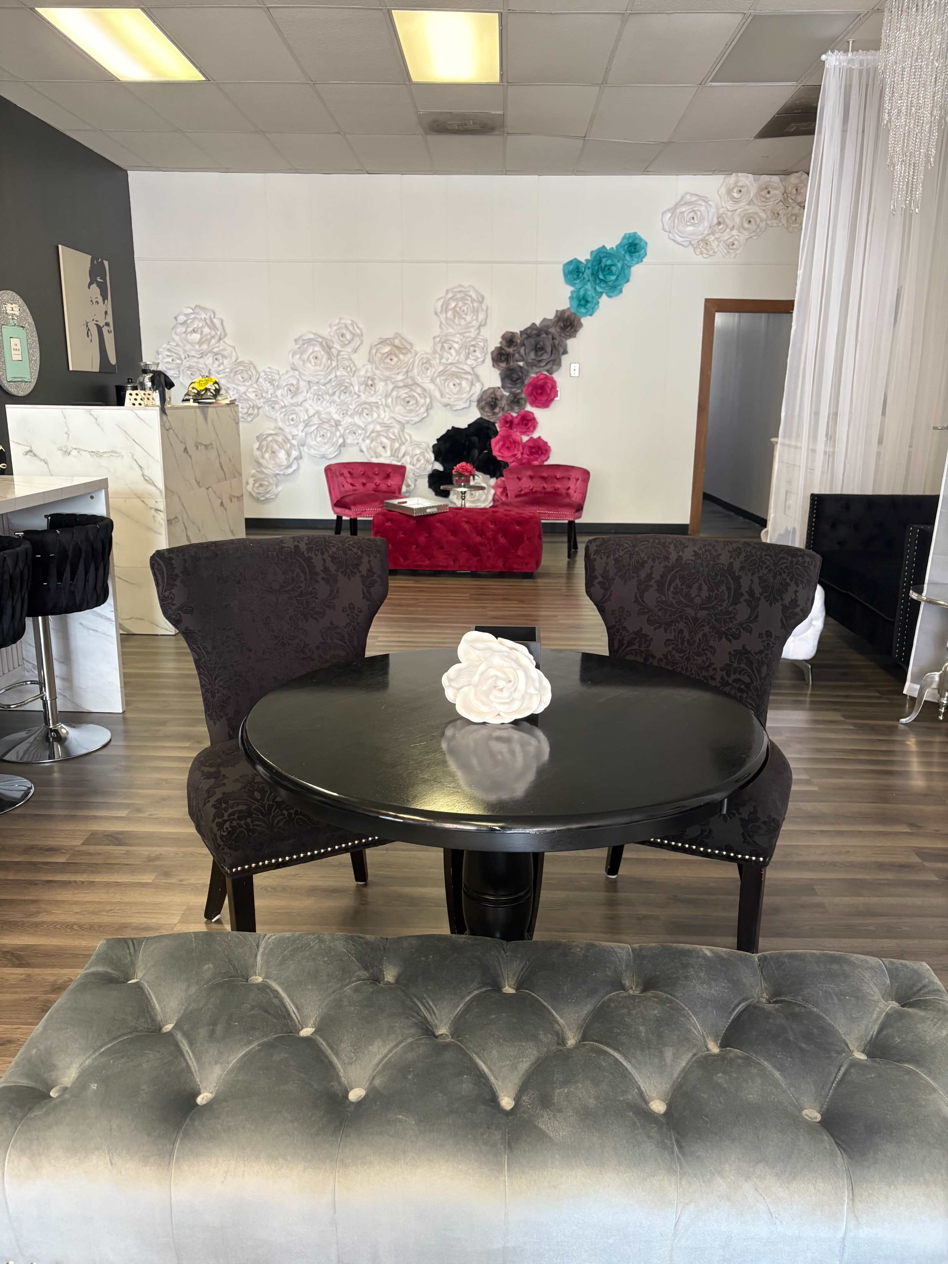 The image shows a modern lounge area featuring a black round table with two ornate chairs, a tufted gray bench, and colorful flower decorations on the wall behind red and black seating.