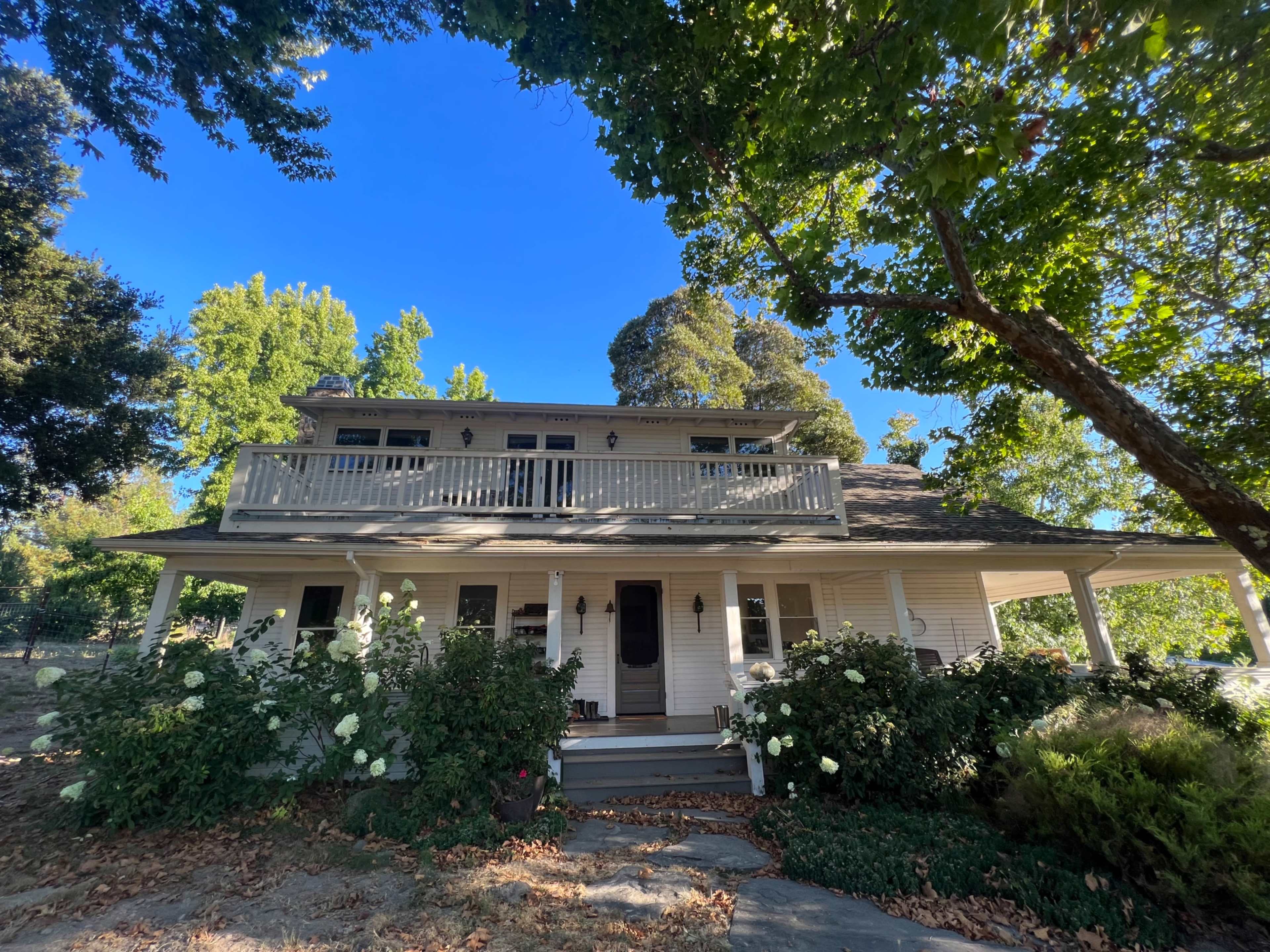 A large, two-story house with a front porch and balcony is surrounded by greenery and flowering bushes under a clear blue sky.