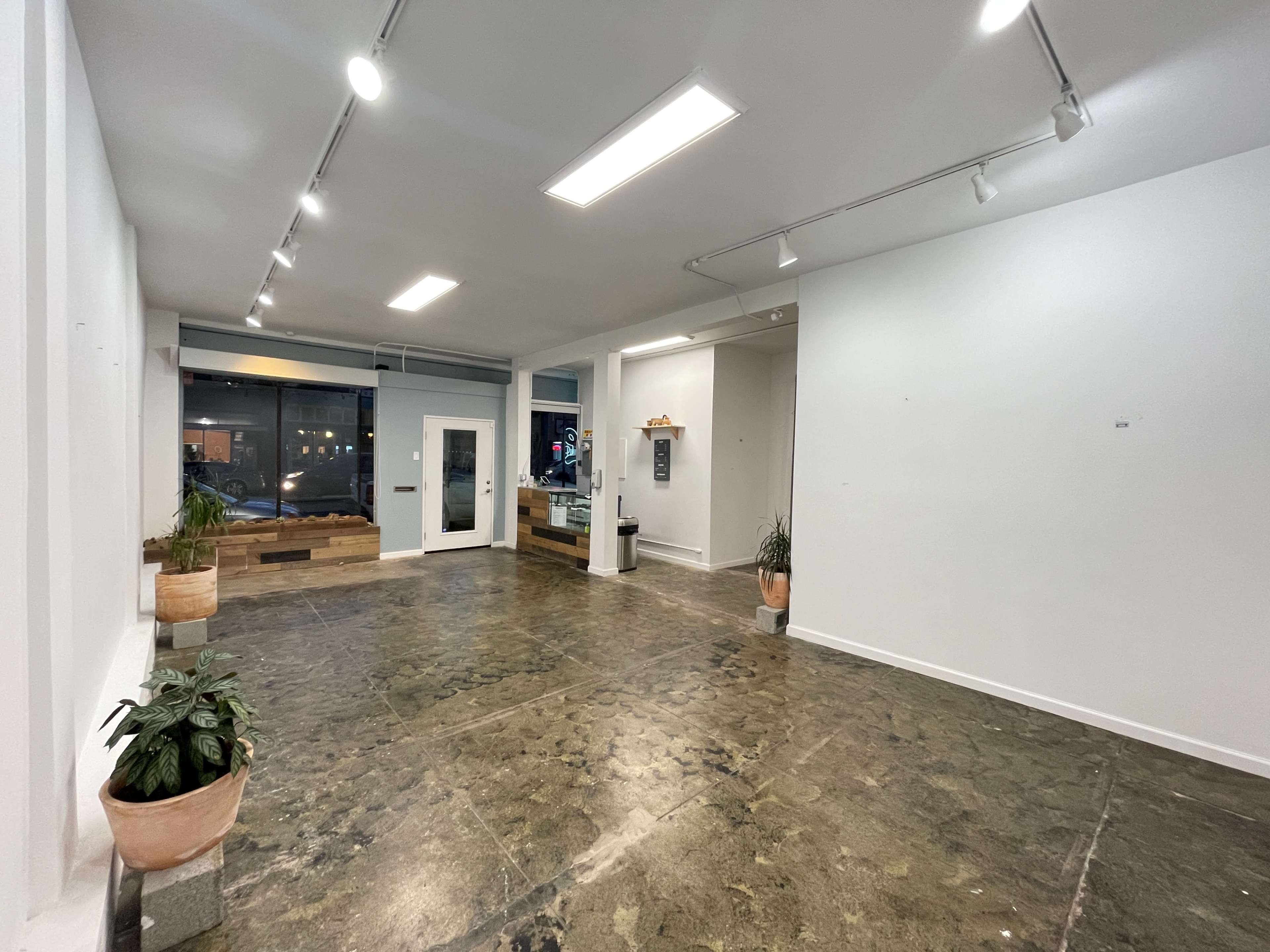 An empty interior space with polished concrete flooring, white walls, and a large window, featuring a door and a service counter in one corner.