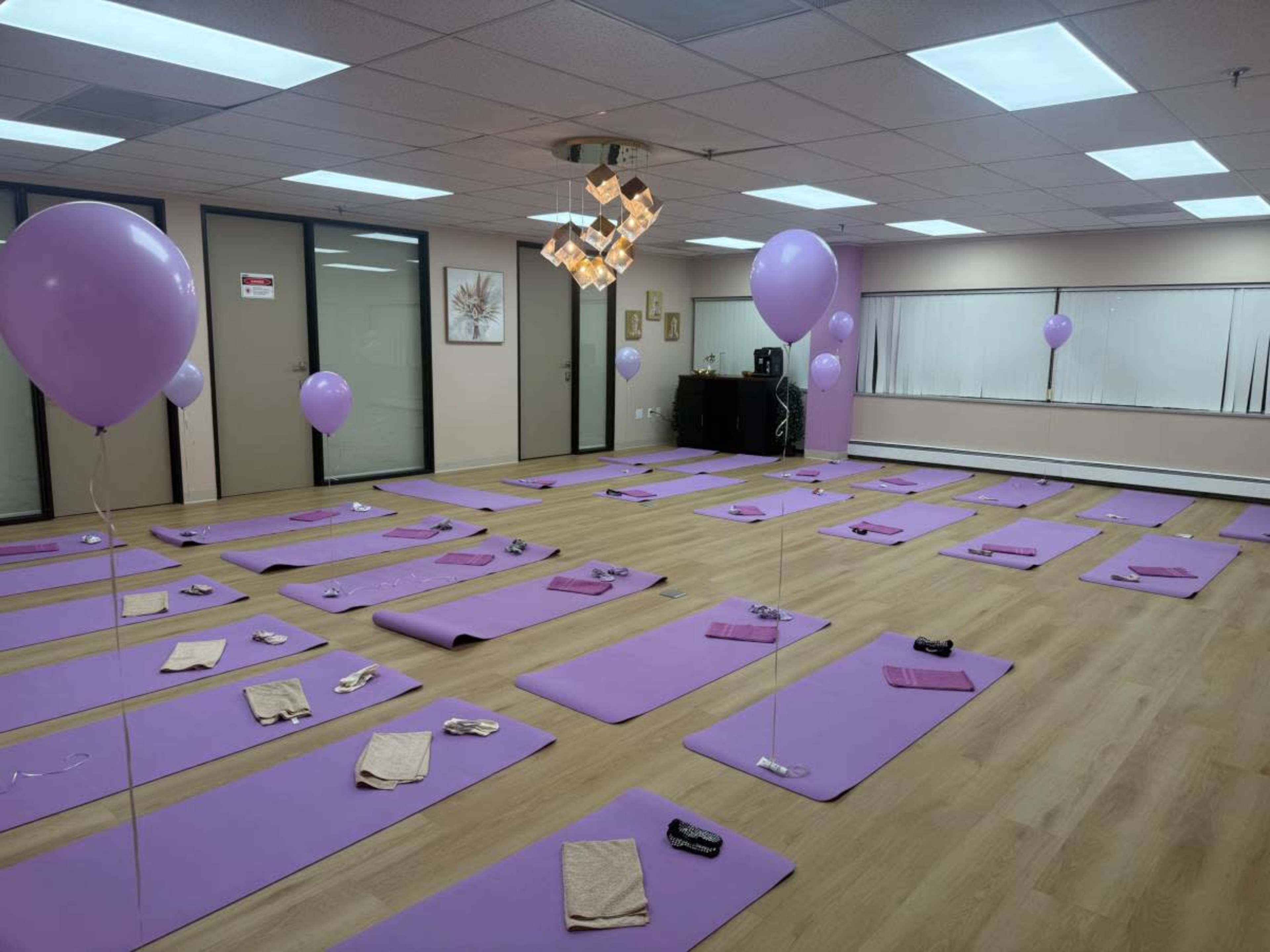 The image shows a spacious room with several purple yoga mats arranged on a wooden floor, accompanied by balloons and towels on each mat.