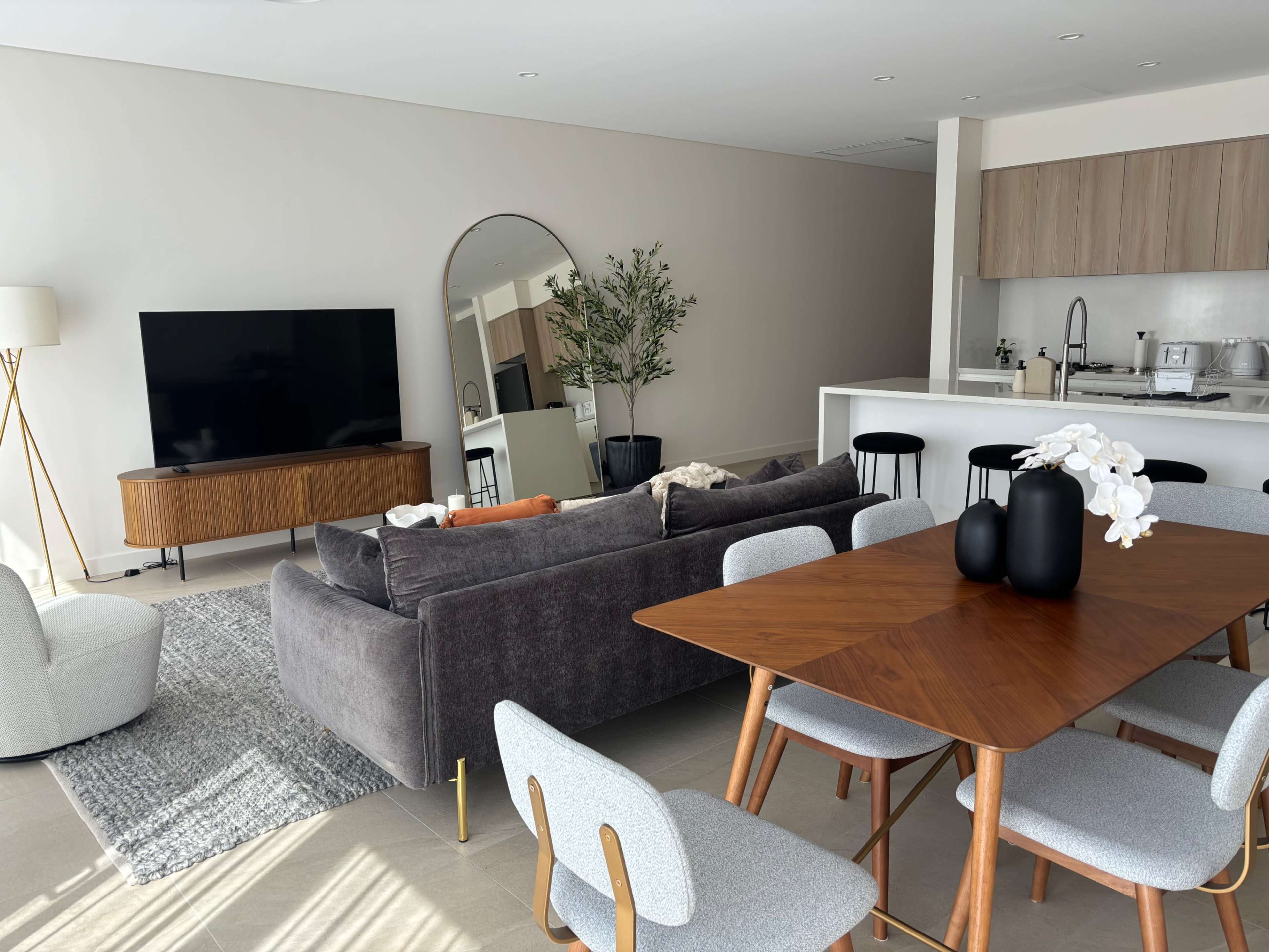 A modern living space featuring a gray sofa, a wooden dining table with chairs, a large television, and a kitchen area in the background.